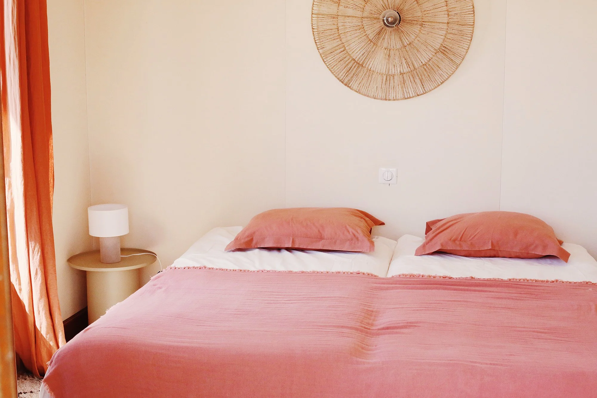 Chambre avec lit double, oreillers rose, couverture rose, lampe de chevet, mur beige, décoration naturelle au plafond, rideaux orange.