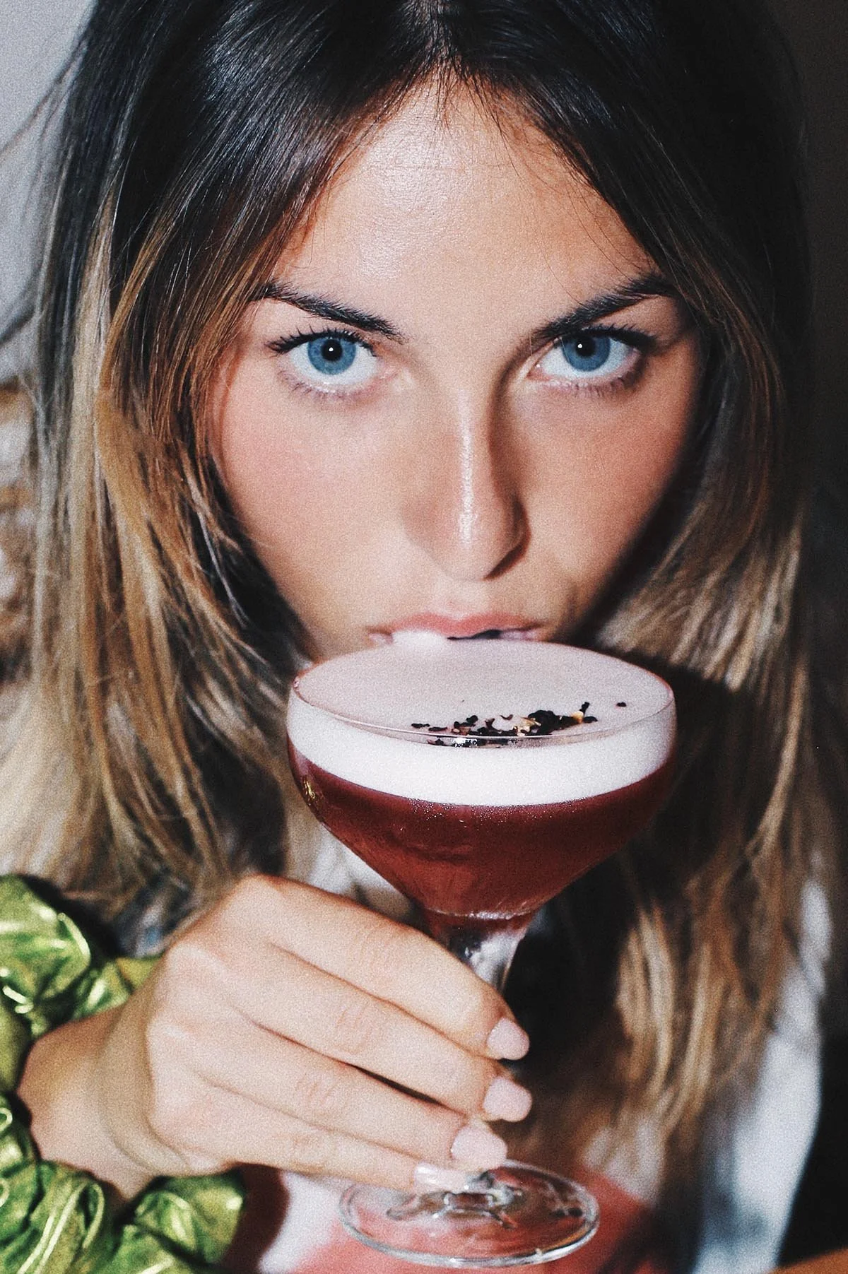 Une femme aux yeux bleus tient un verre à cocktail rouge avec une mousse blanche et des garnitures noires, regardant directement l'objectif.
