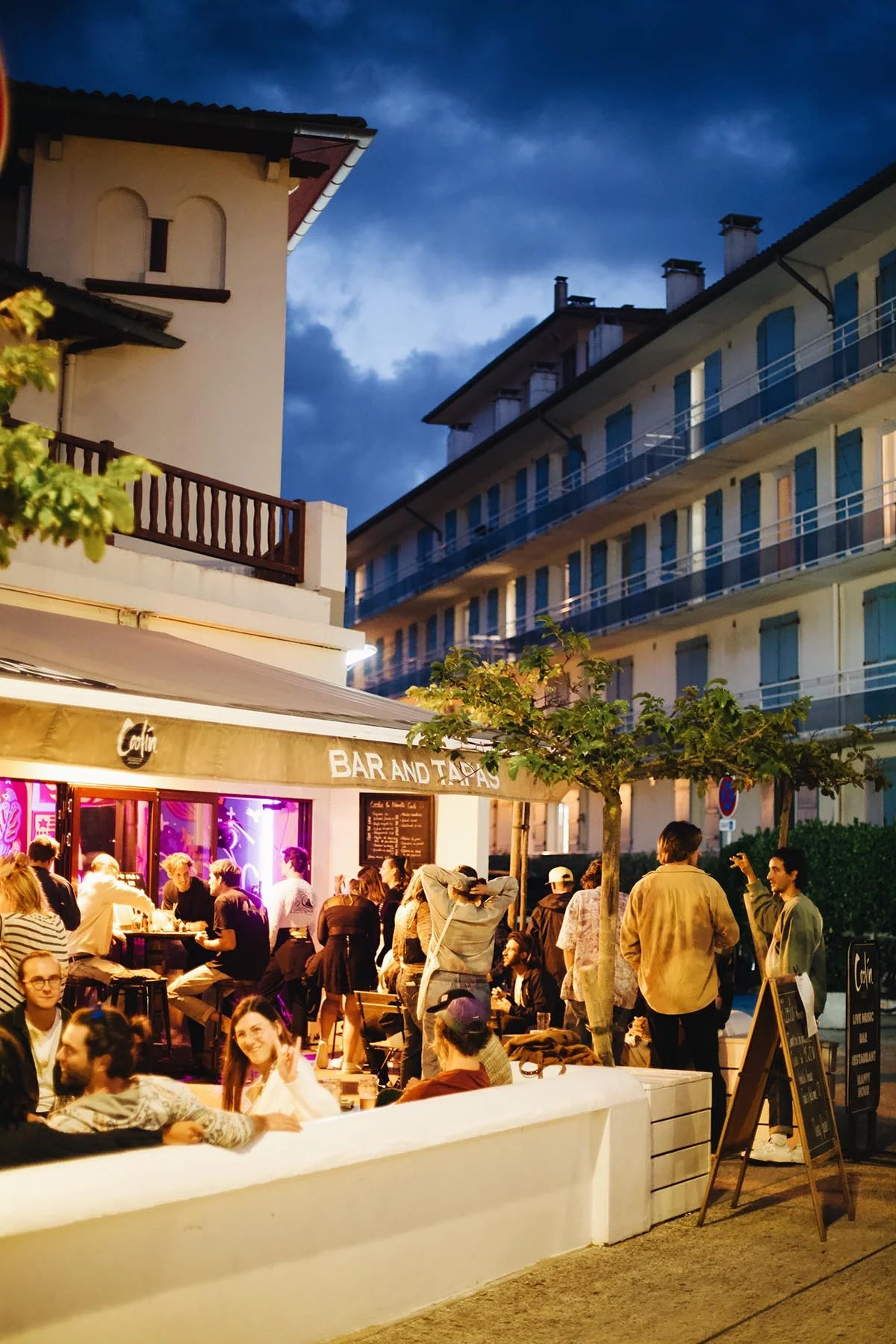 Une scène nocturne devant un bar avec des gens assis et discuter, des arbres et un bâtiment résidentiel en arrière-plan, ciel nuageux.