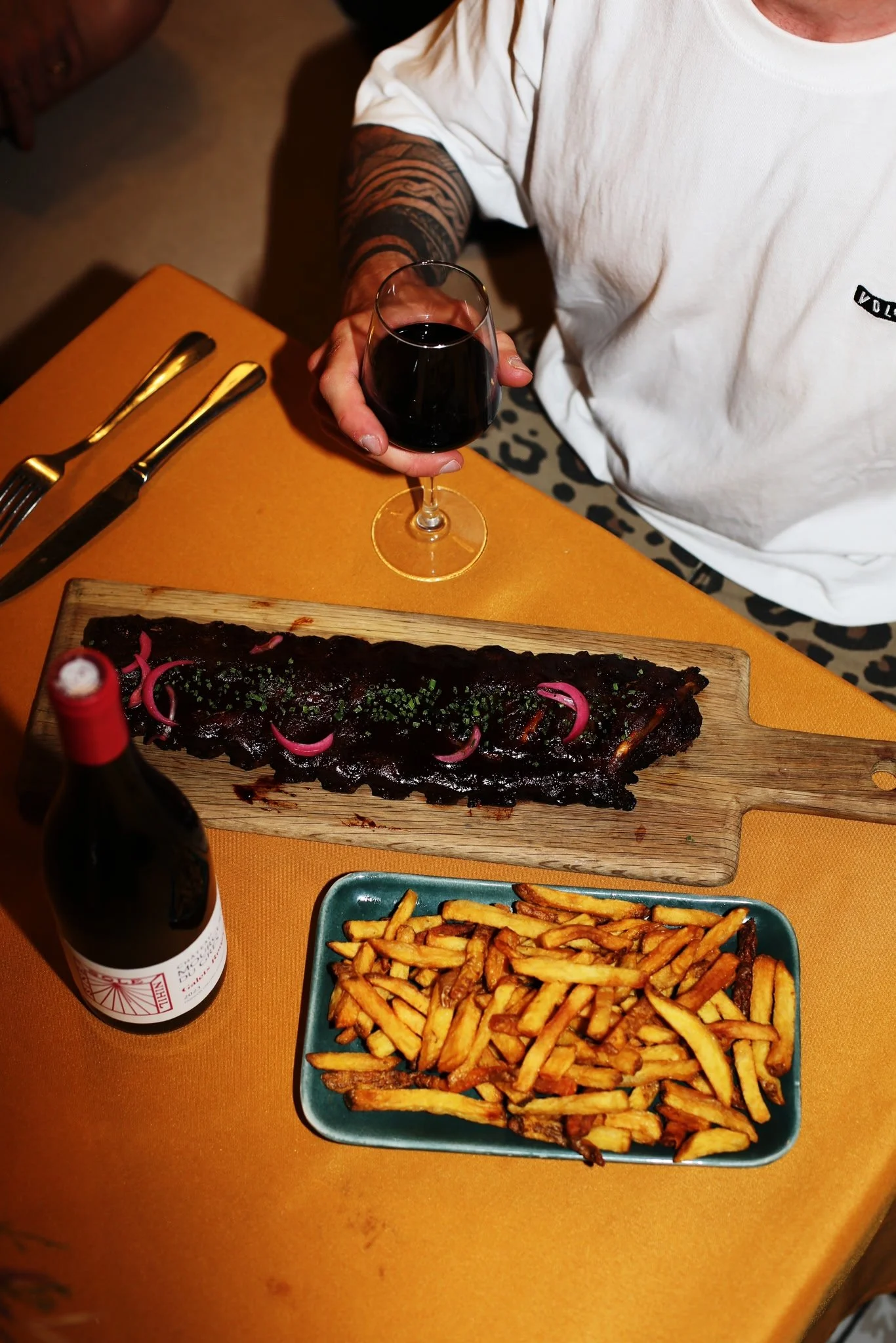 Une personne tenant un verre de vin rouge assise à une table avec une assiette de ribs, un plat de frites, et une bouteille de vin dans un restaurant.