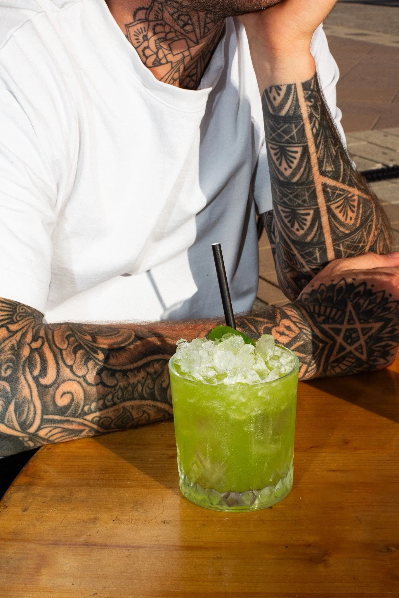 Une personne tatouée aux bras et au cou est assise à une table en bois. Elle porte un t-shirt blanc et une montre noire. Devant elle se trouve un verre vert rempli de glaçons et un cocktail vert vif, accompagné d'une paille.