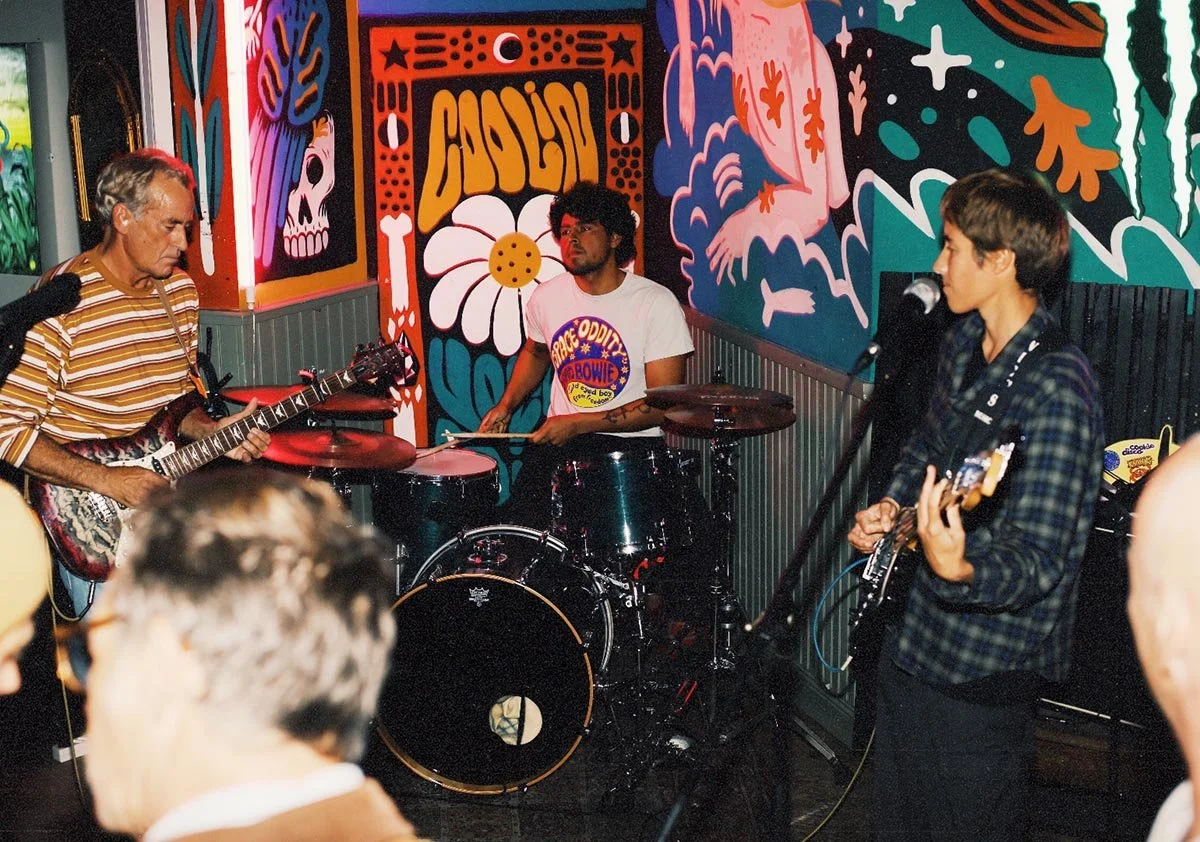 Groupe de trois musiciens jouant dans un espace décoré avec des murs colorés et un mur avec des dessins de fleurs, de crânes et d'autres motifs artistiques.