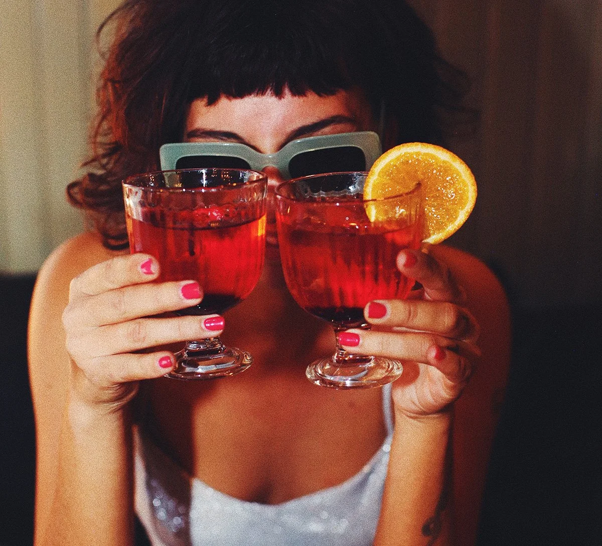 Une femme portant des lunettes de soleil tient deux verres de cocktail rouge avec une tranche d'orange, souriant derrière les verres.