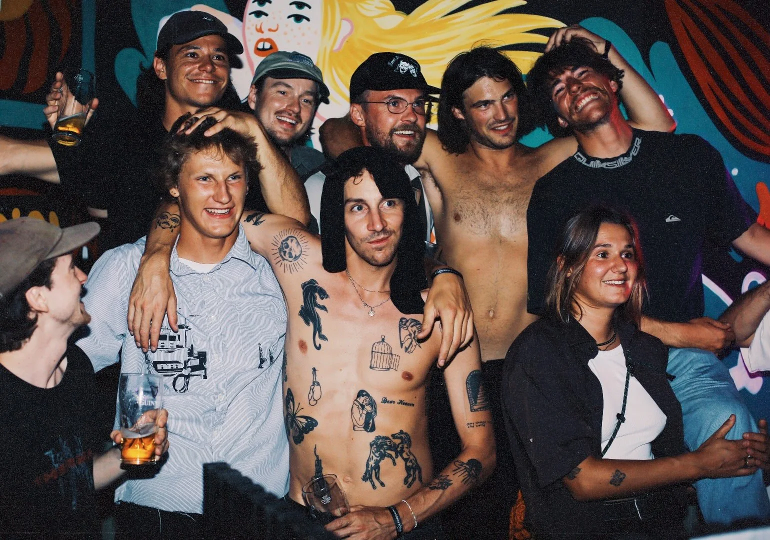 Groupe de jeunes hommes et femmes souriants posant ensemble lors d'une fête ou d'un rassemblement, certains en tenue décontractée ou torse nu, avec tatouages visibles, fond coloré.