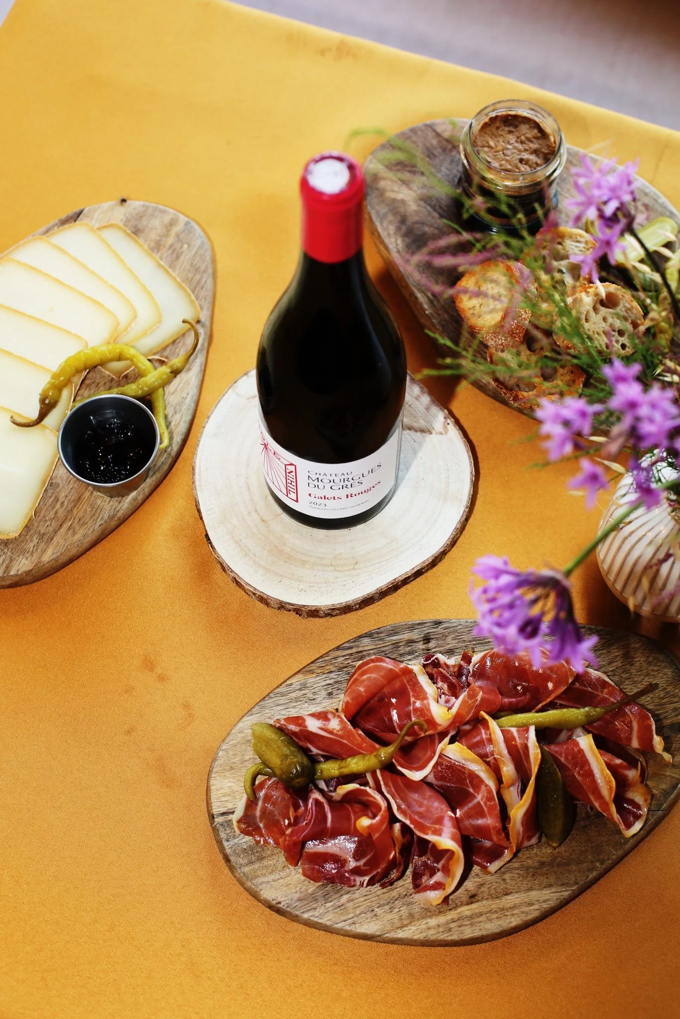 Plateau de charcuterie avec jambon cru, cornichons, et fromage, accompagnée d'un verre de vin rouge et d'une jarrette de pâté, sur fond orange avec des fleurs violettes