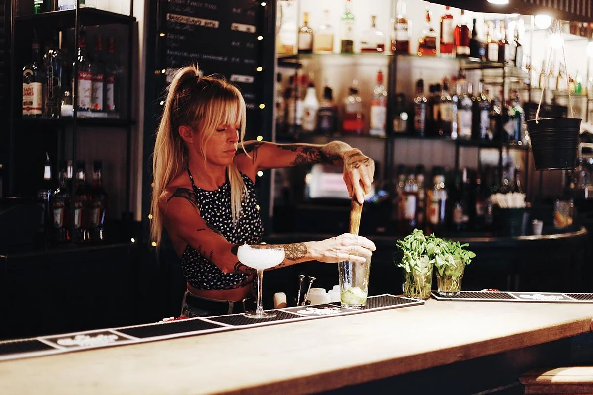 Une femme barista prépare un cocktail derrière un comptoir de bar, avec des bouteilles d'alcool en arrière-plan et des herbes fraîches devant elle.