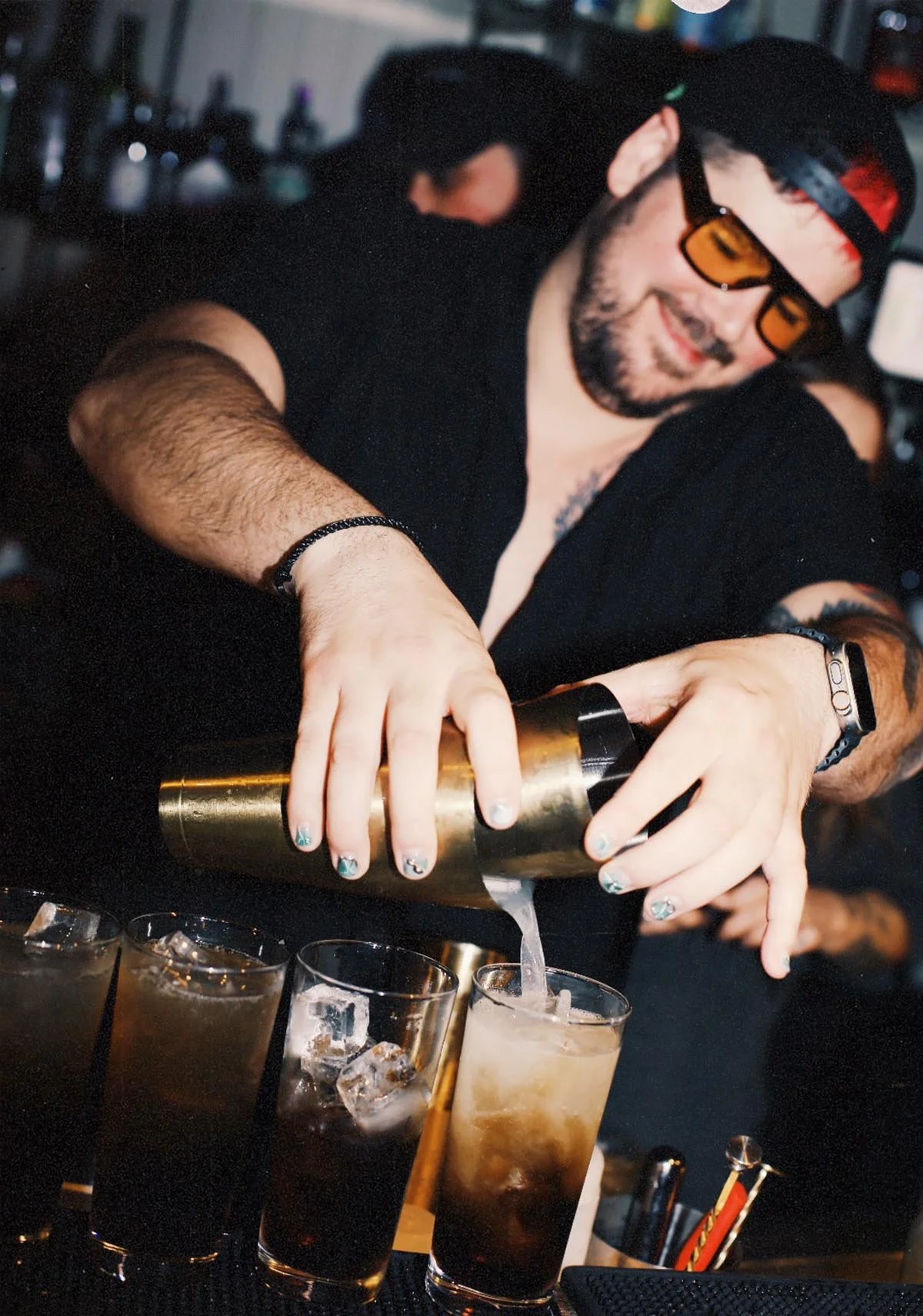Un homme souriant, portant des lunettes de soleil orange, un bonnet noir, et une barbe, sert des cocktails dans un bar.