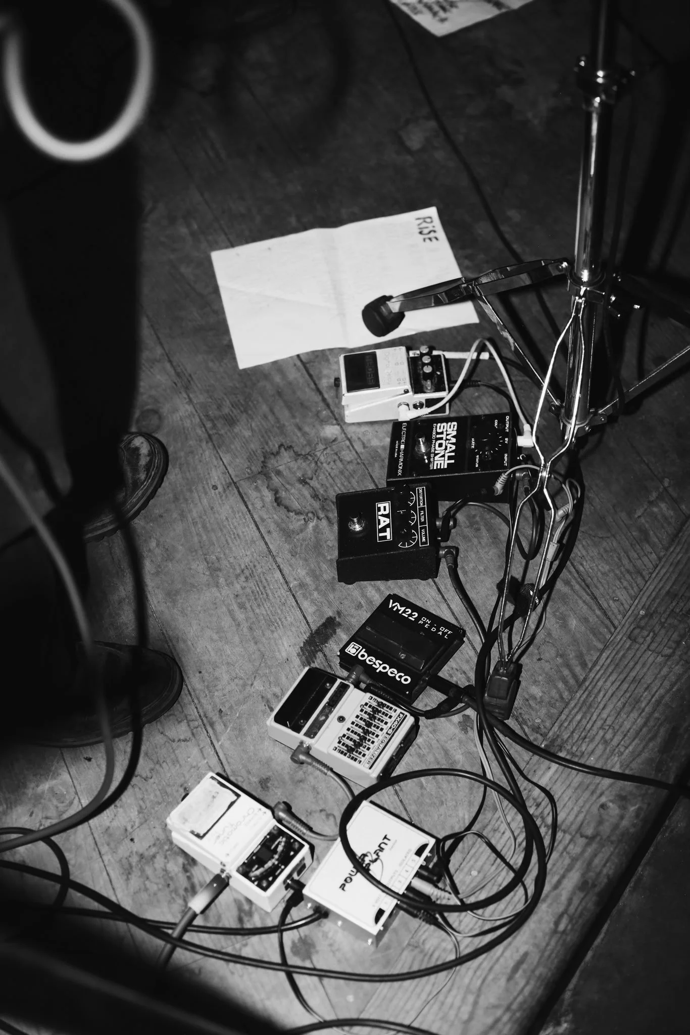 Une scène de musique avec plusieurs pédales d'effets, un métronome, un pied de support et un papier avec l'inscription 'Rêve' sur un plancher en bois. L'image est en noir et blanc.
