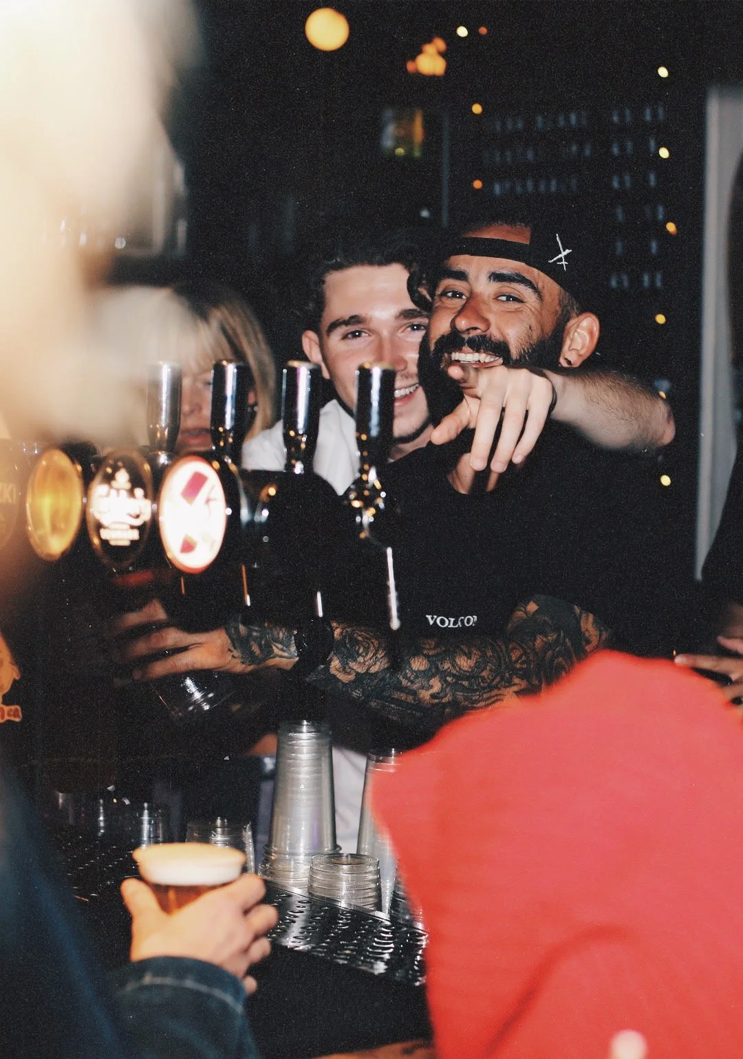 Groupe de personnes souriantes faisant un toast dans un bar ou club nocturne, avec des verres à bière en arrière-plan et des lumières ambiance.