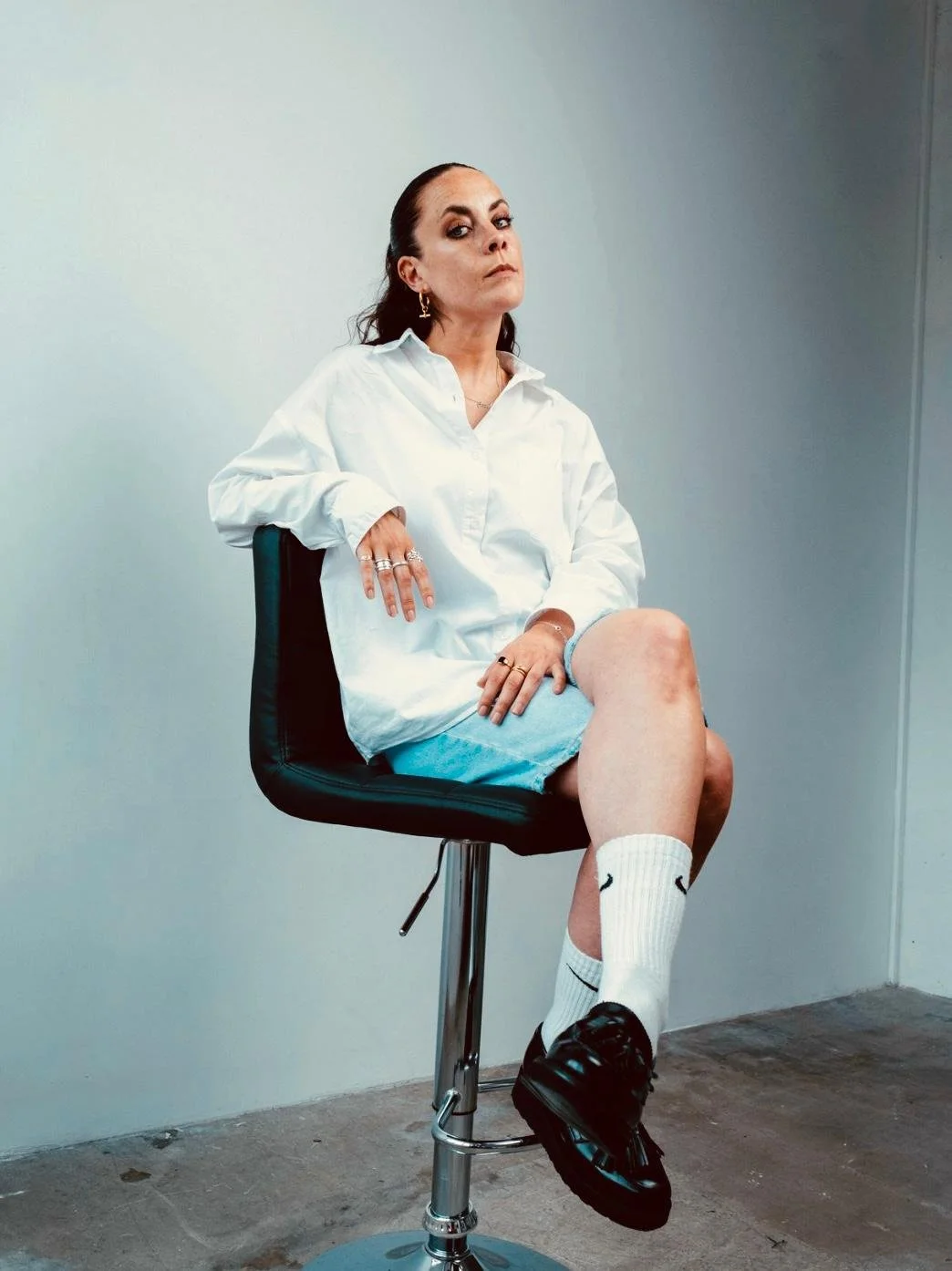 A woman with dark hair pulled back, wearing a white oversized shirt, light blue shorts, white Nike crew socks, and black shoes, sitting on a black office chair against a plain wall.