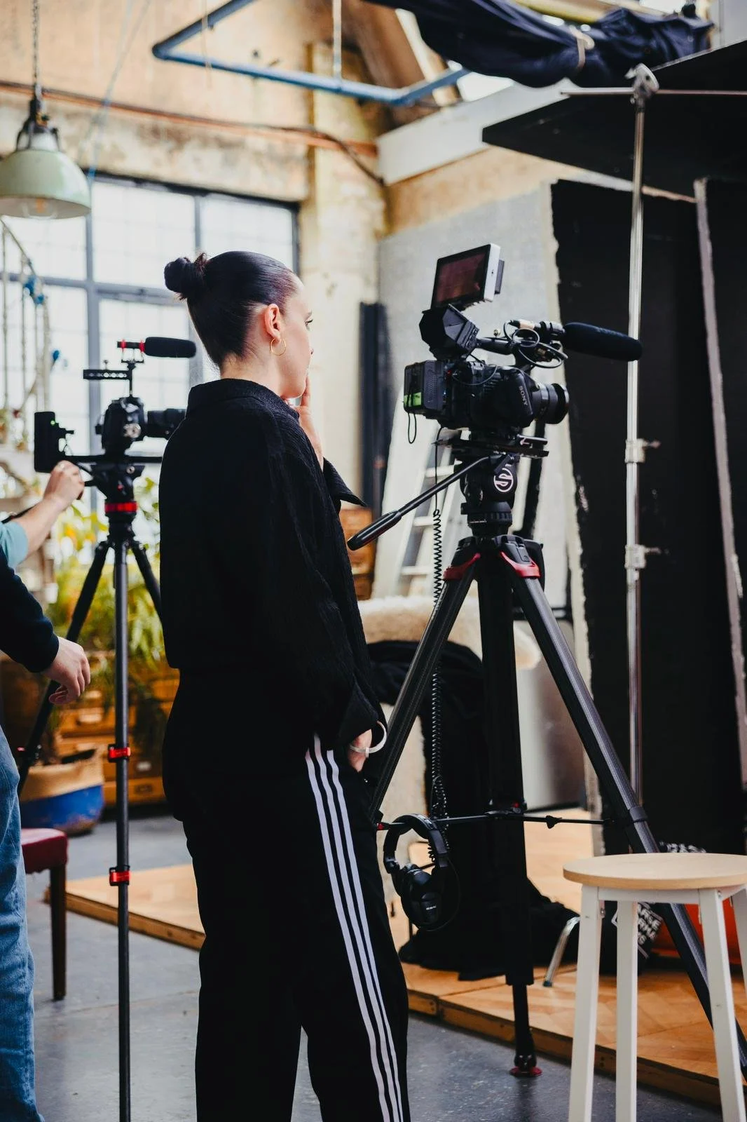 A woman with dark hair in a bun observing a professional video camera setup in a studio with natural light, brick walls, and studio equipment.