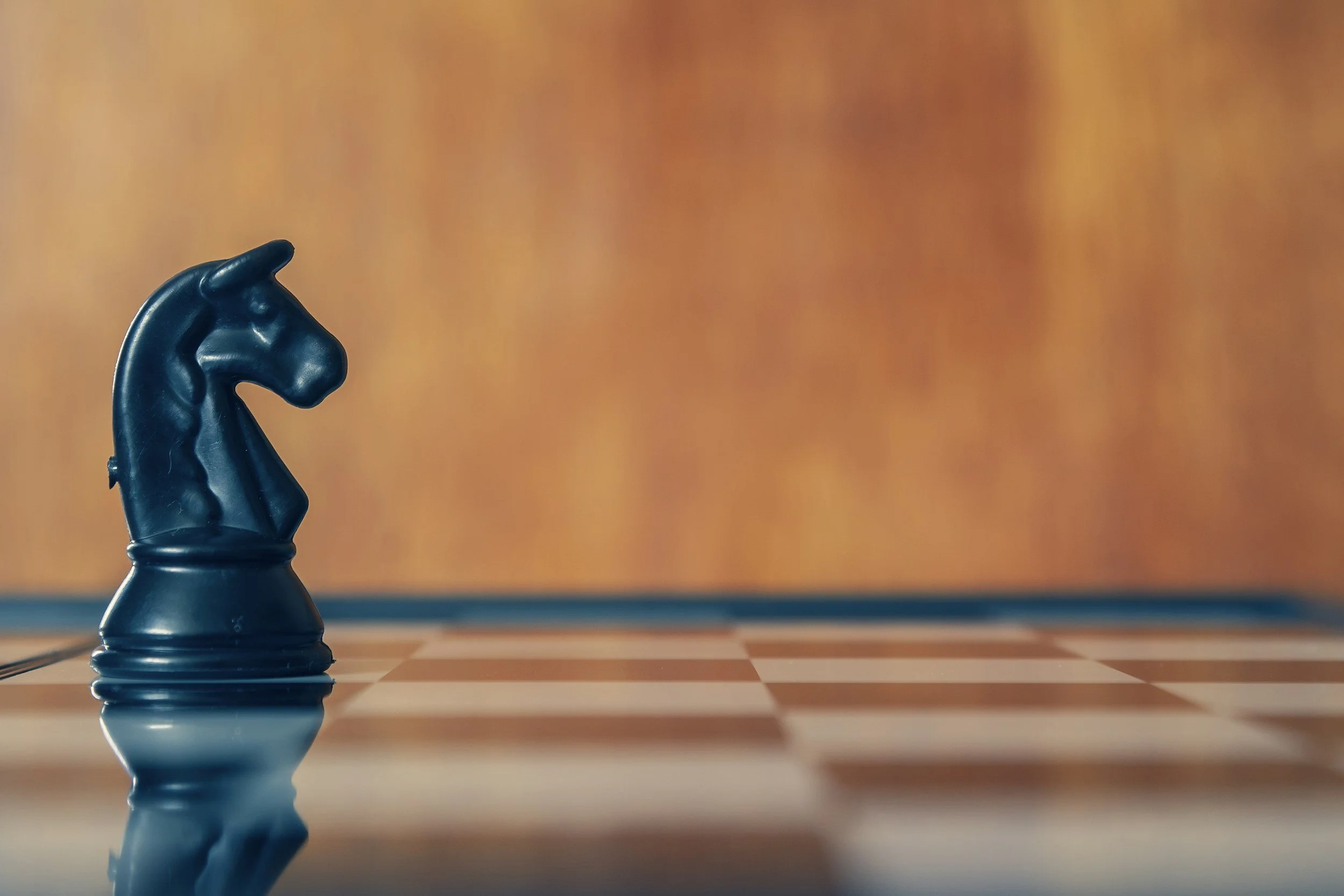 A close-up view of a black king chess piece on a chessboard, with the piece's reflection visible on the polished surface below. The background is blurry with warm orange tones.