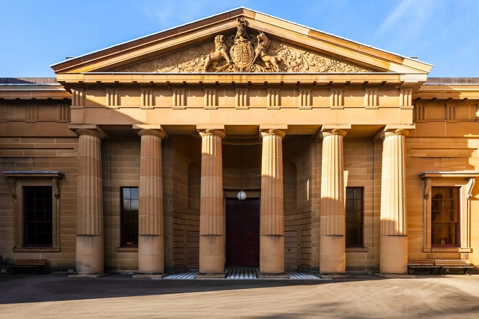 Front view of a neoclassical building with six large pillars, a triangular pediment with intricate carvings, and a coat of arms with two lions at the top. The building is made of beige stone, with windows and sunlight casting shadows.