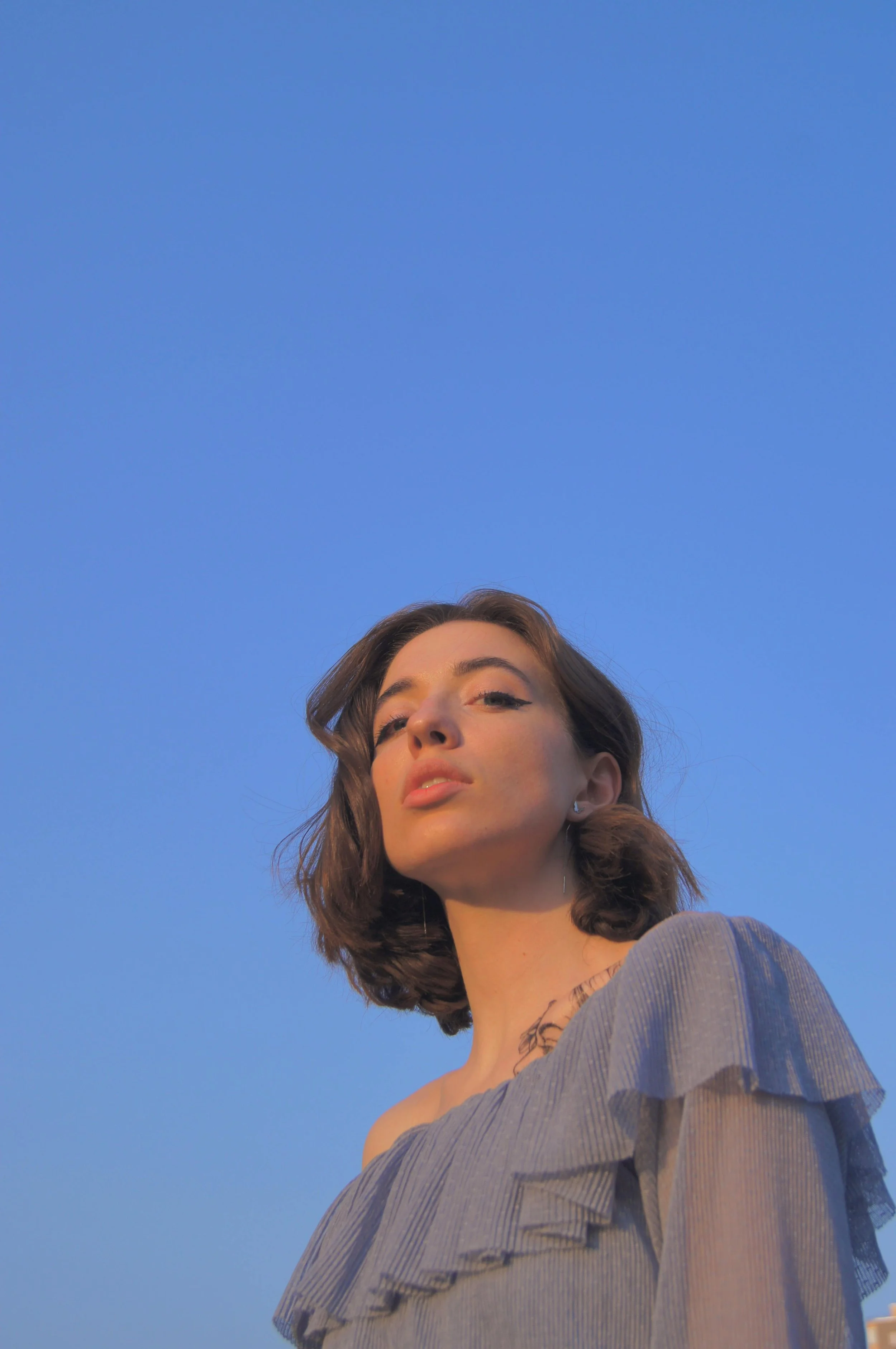 A woman with shoulder-length brown hair and light skin is looking slightly upward with a neutral expression. She is wearing a gray off-the-shoulder top and standing outdoors against a clear blue sky.