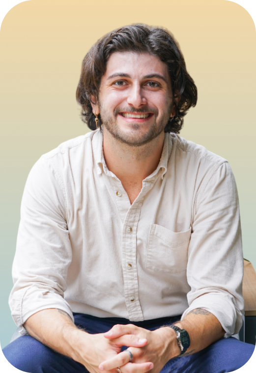 A smiling man with dark hair, beard, and earrings, wearing a beige button-up shirt and dark pants, sitting against a blurred yellow and green background.