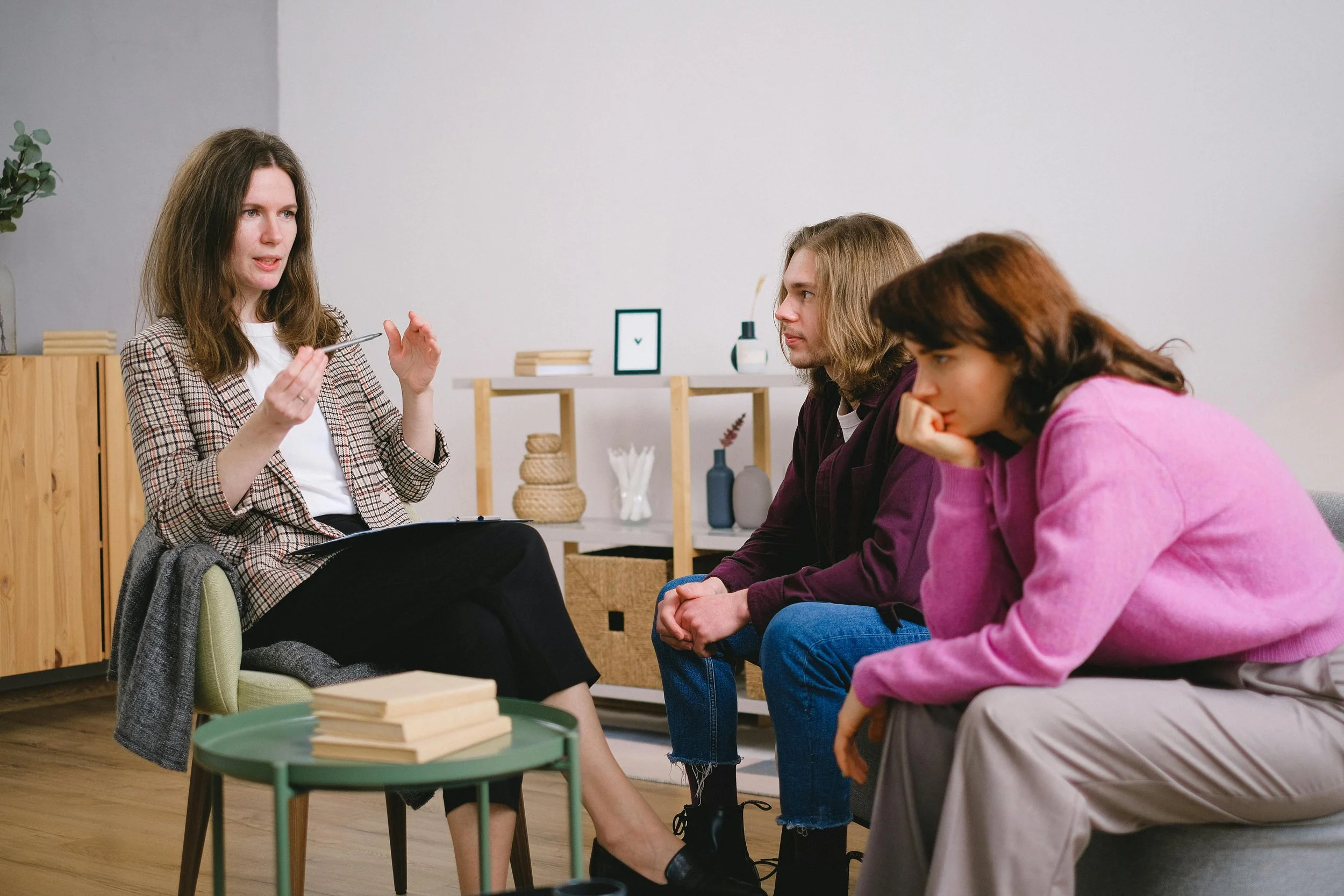 A woman leading a discussion with three young adults in a modern, minimalistic room. The woman is gesturing with her hands, while the others listen attentively. The room has a small table with books and decorative items and a plant in the background.