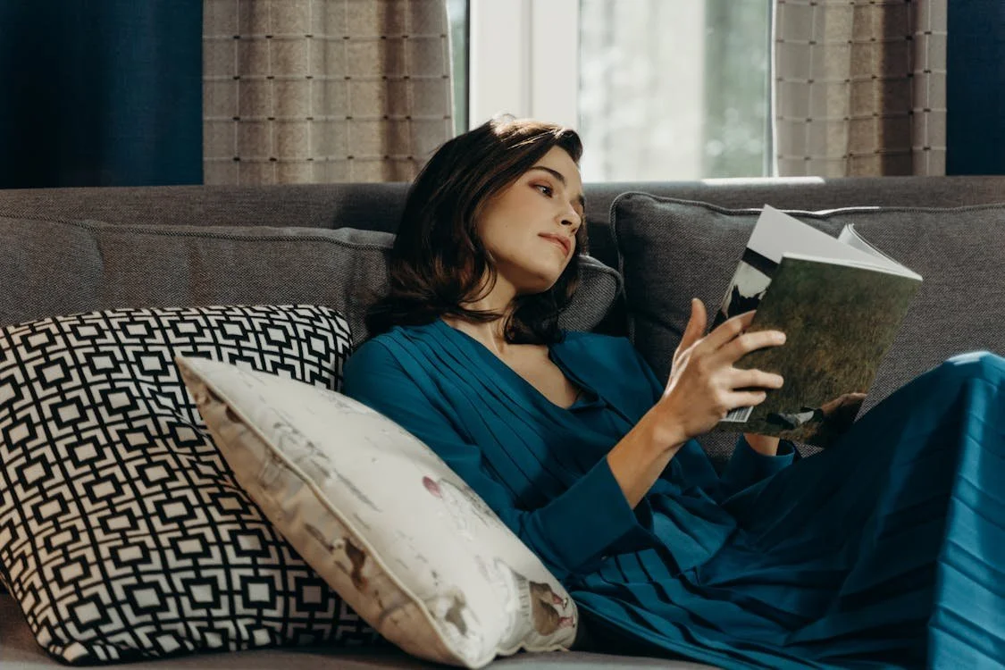 A woman with dark hair in a blue dress reclining on a sofa, reading a book, with decorative pillows and a window in the background.