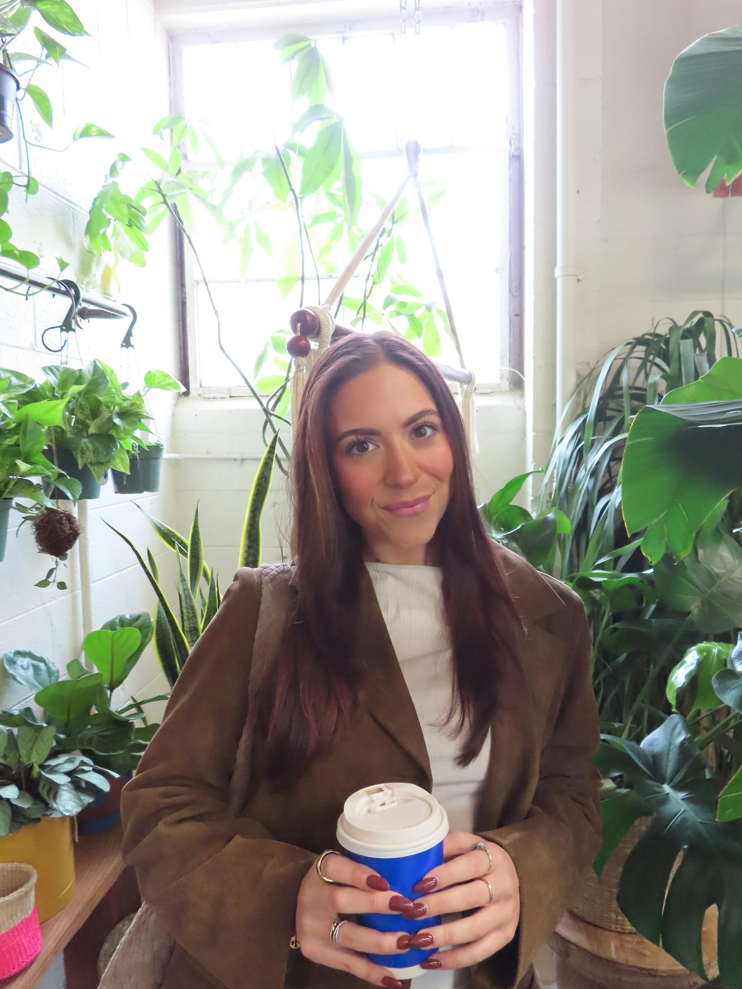 A woman with long reddish-brown hair, holding a disposable coffee cup with a blue sleeve, standing amidst lush green indoor plants in a bright room.