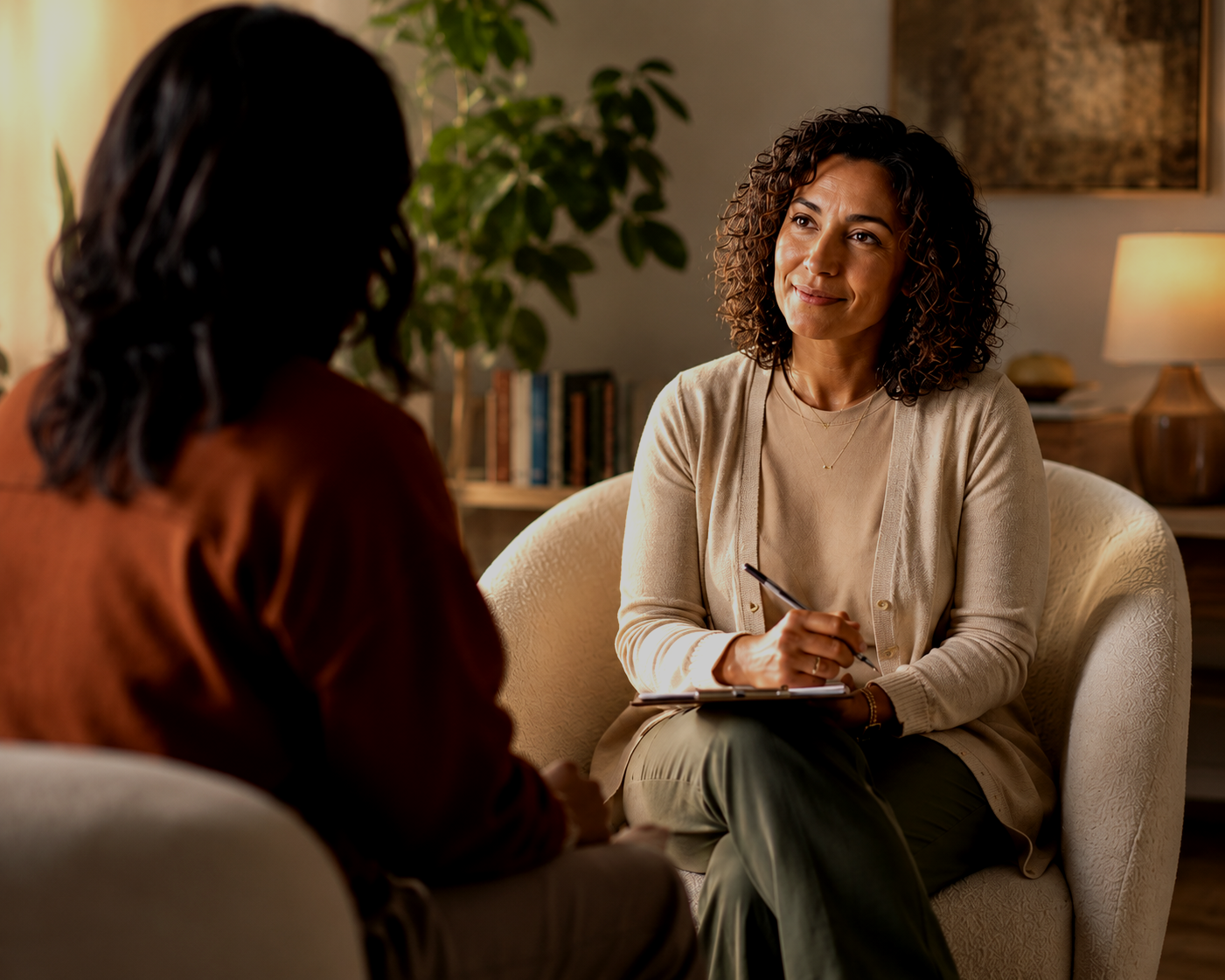 A woman with curly hair and a beige cardigan smiling during a conversation with another woman in a cozy, well-lit room.