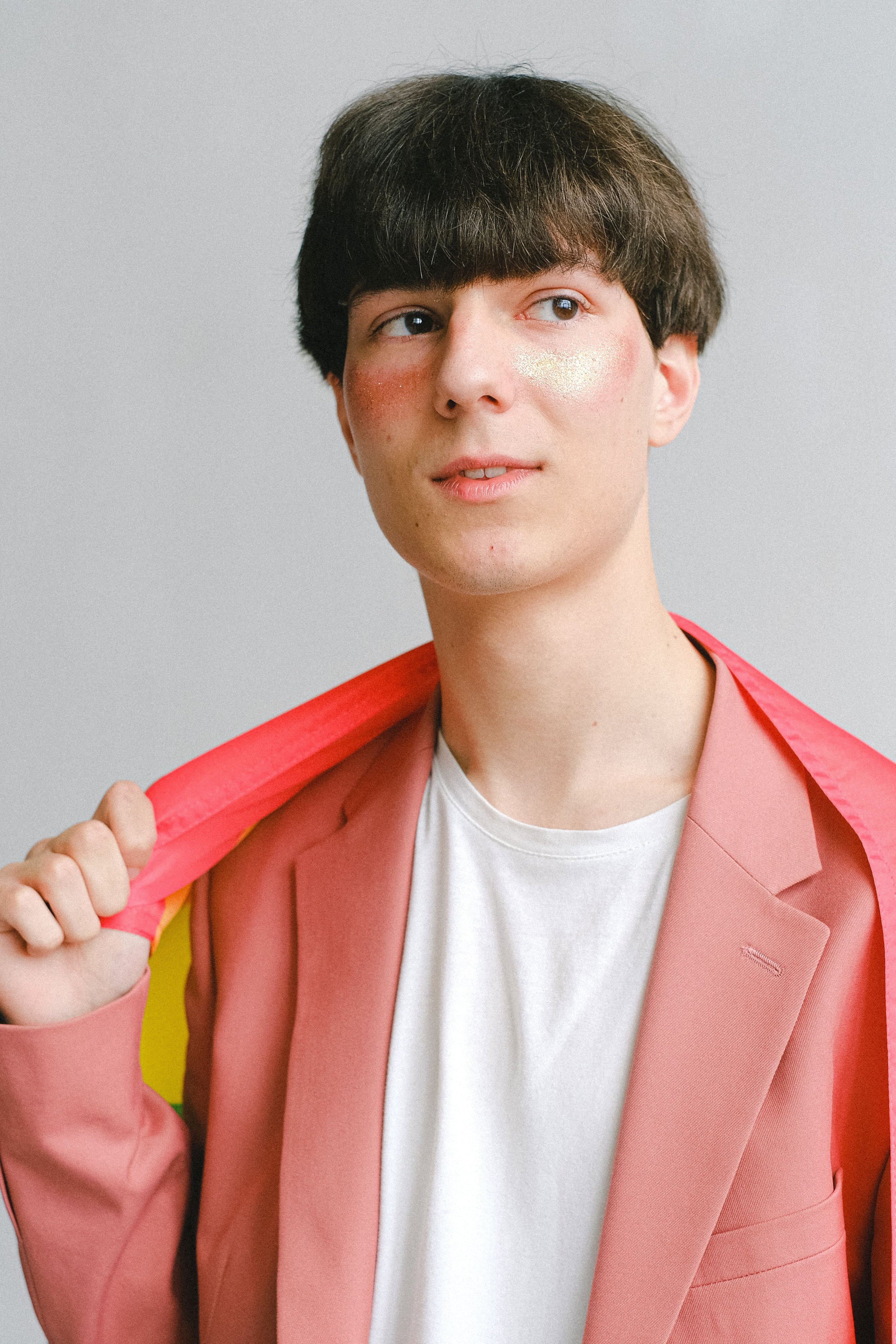 A person with short dark hair wears a pink blazer over a white shirt, holding a pink and yellow object, standing against a plain light grey background.