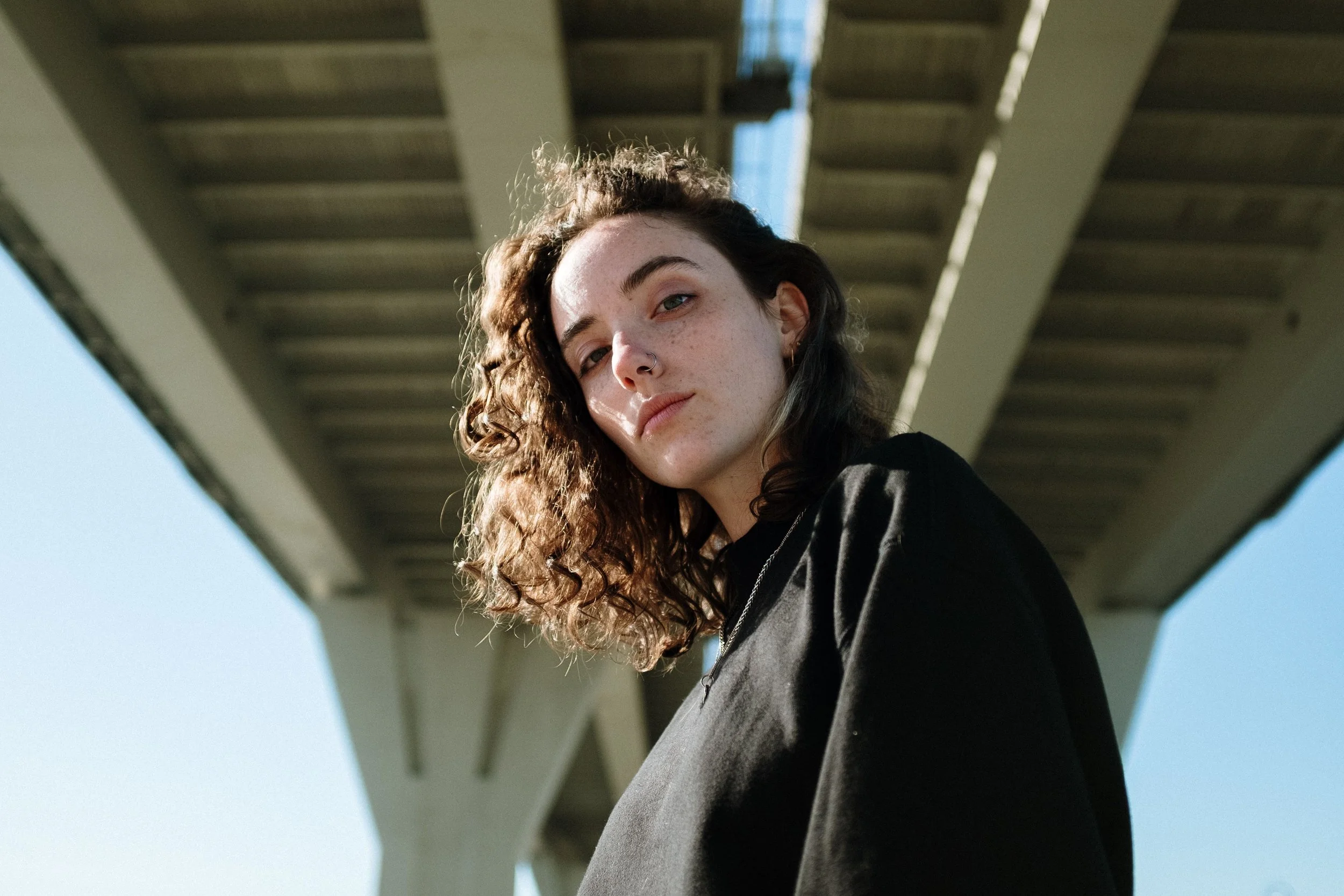 A queer person with curly hair stands confidently under a contemporary bridge.