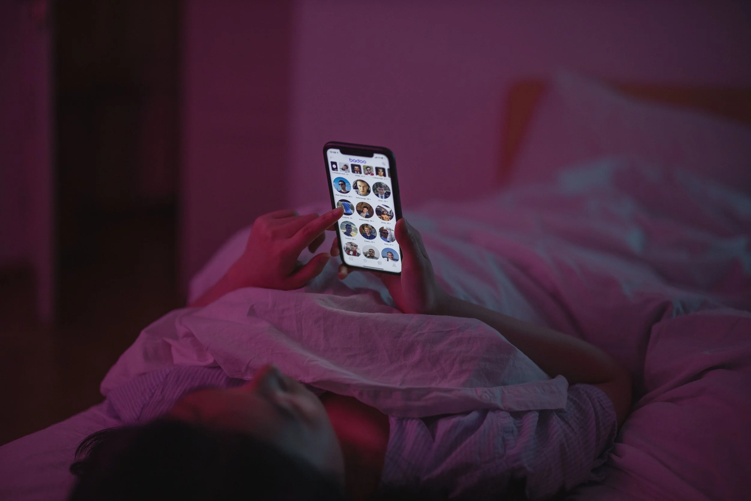 A woman relaxes in bed at night, browsing on her smartphone under pink ambient lighting.