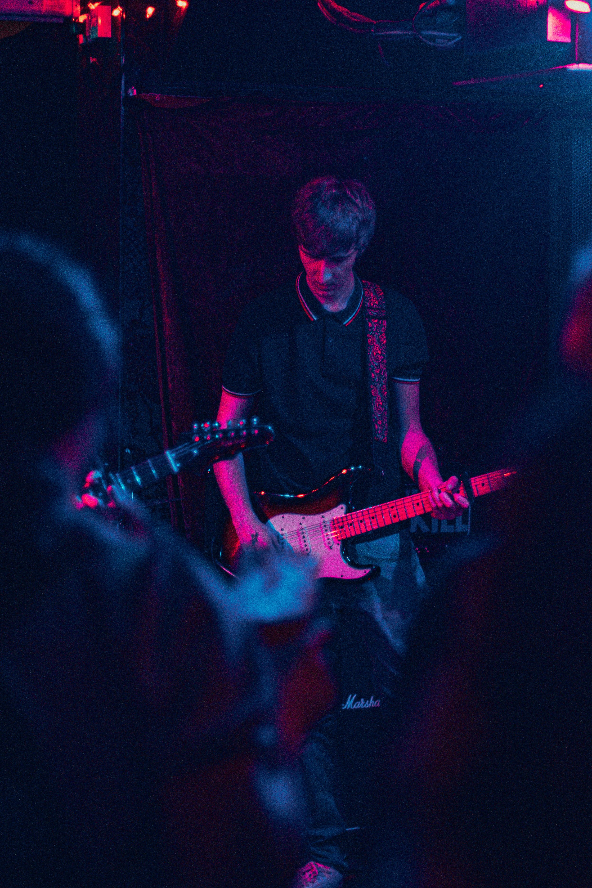 A young man playing an electric guitar on stage in a dimly lit, vibrant environment with pink and purple lighting.