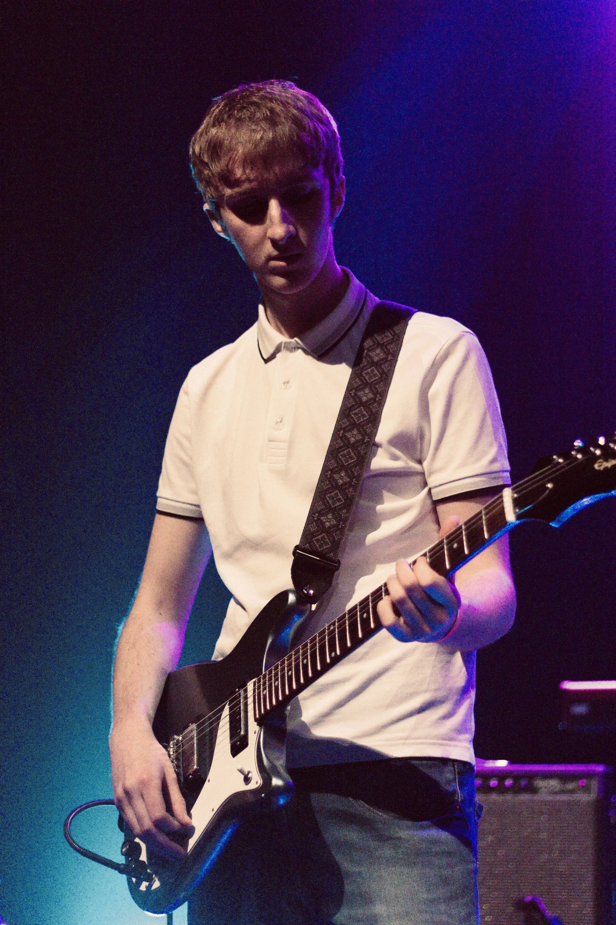 A young man with short light brown hair and playing an electric guitar on stage with purple and blue lighting.