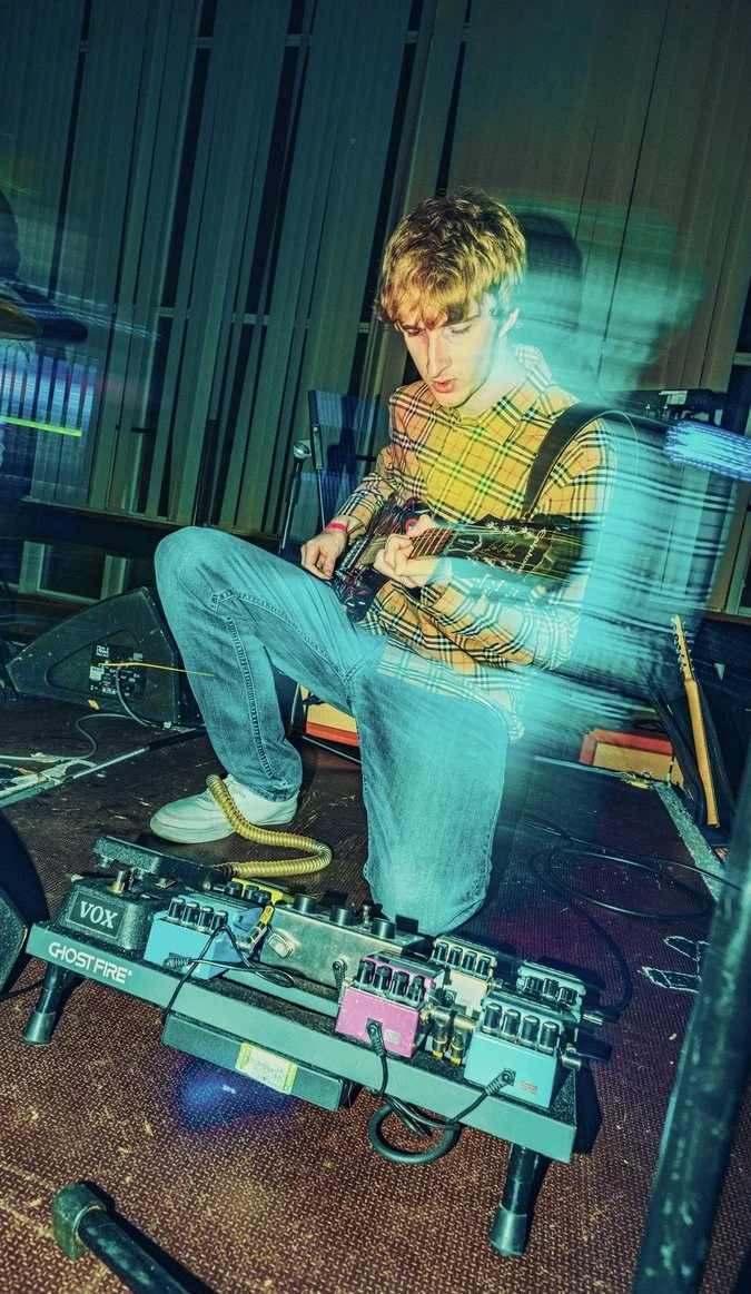 A young man wearing a yellow plaid shirt and blue jeans is sitting on a stage, playing an electric guitar. There are guitar effects pedals and electronic equipment in front of him. The background includes a wooden wall with vertical panels, and colorful light trails streak across the image, suggesting a dynamic performance or creative environment.