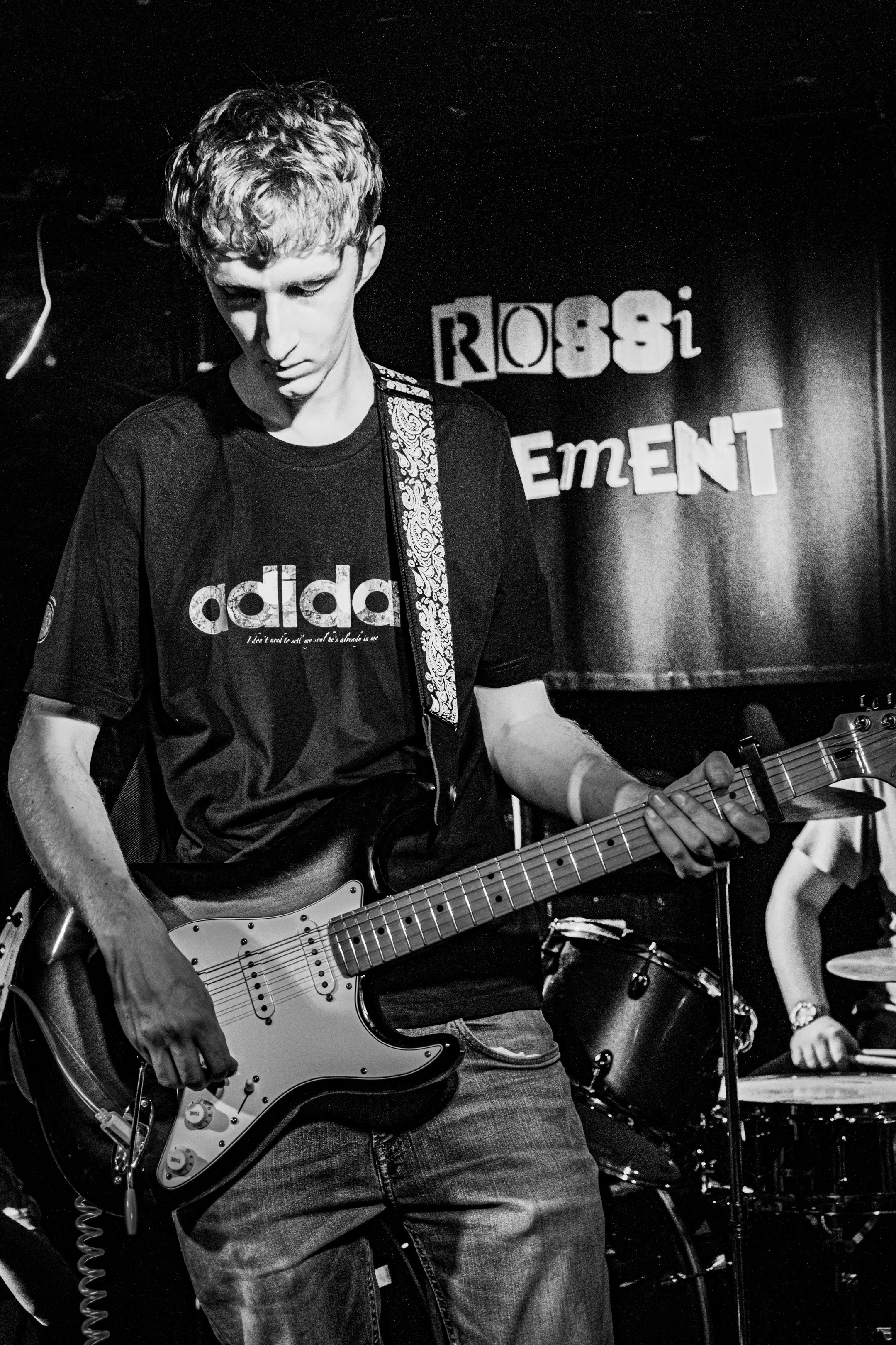 A young male guitarist playing an electric guitar during a live performance at a venue called Rossi bar, with drums in the background, wearing a Adidas T-shirt and jeans.