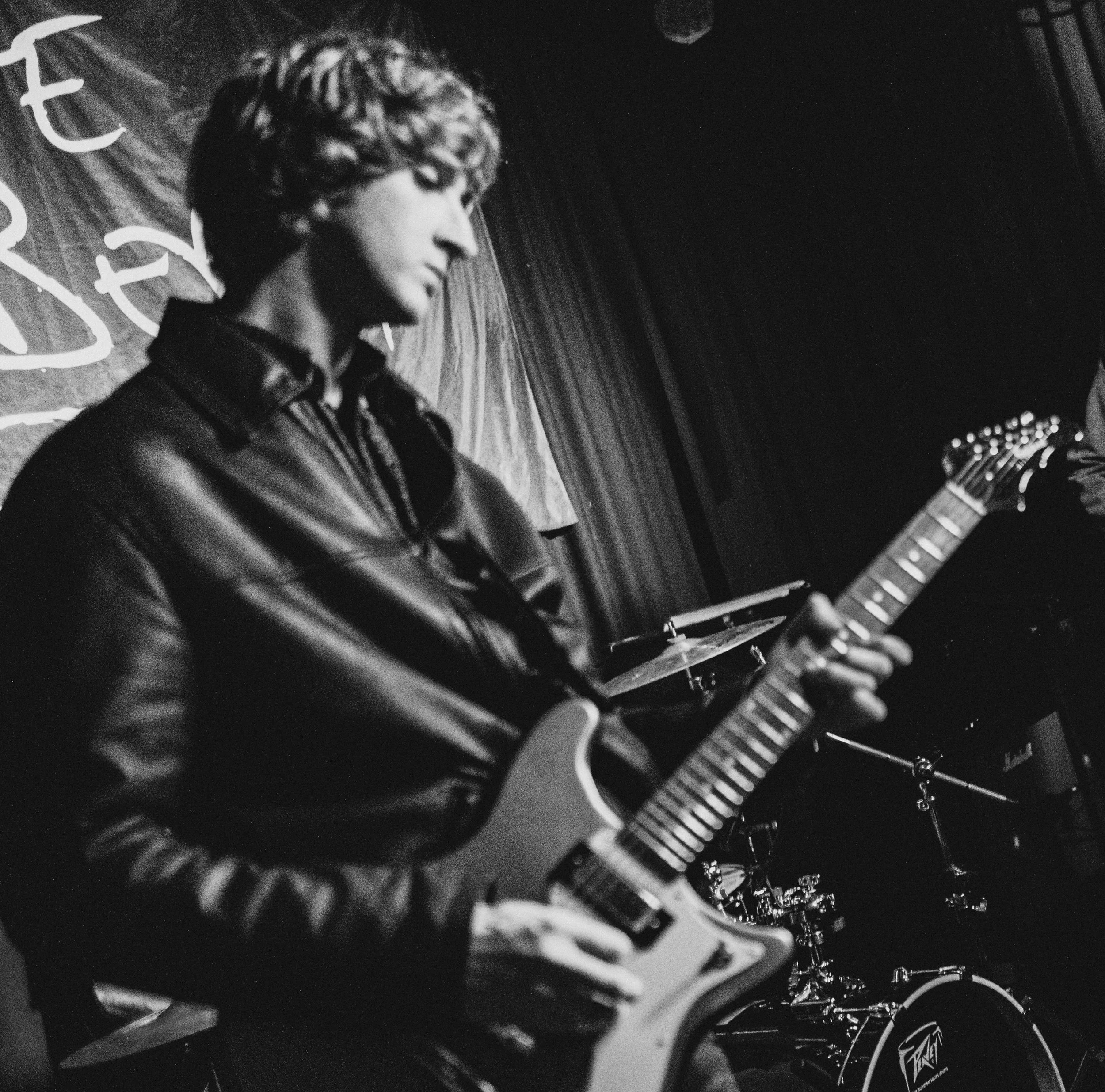 A black-and-white photo of a person playing an electric guitar on a stage. The person has long hair and is wearing a jacket, focused on playing the guitar. The background features a wooden wall and some abstract writing or signs.