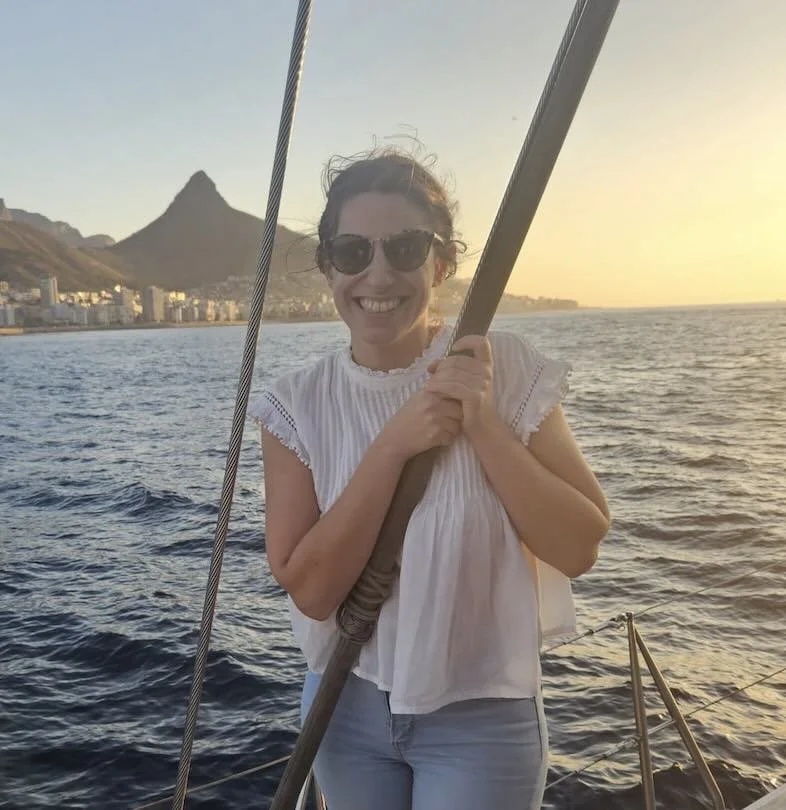 Woman smiling and holding onto a sailboat's rigging on the water at sunset with mountains and a city skyline in the background.