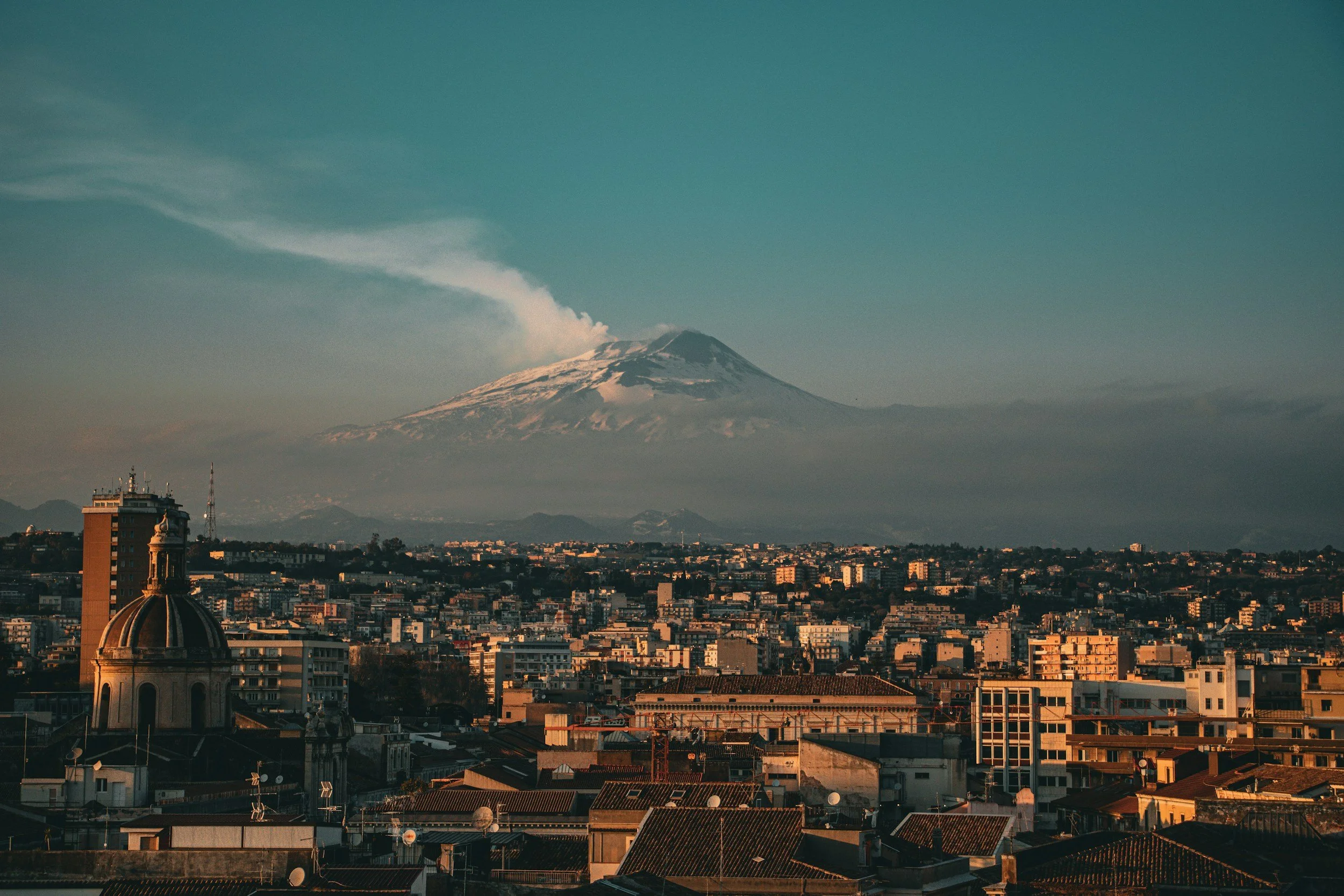 City skyline with buildings and a dome in the foreground, Mount Fuji with snow on its peak and a plume of smoke or steam rising from the top in the background under a blue sky.