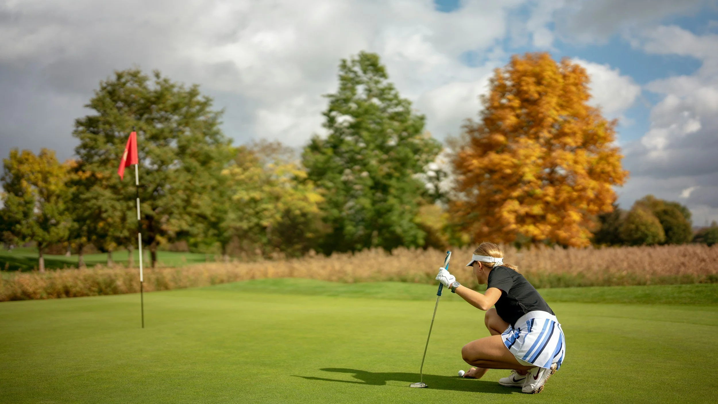 A person crouching on a golf course near the hole, preparing to putt a golf ball with autumn trees in the background.