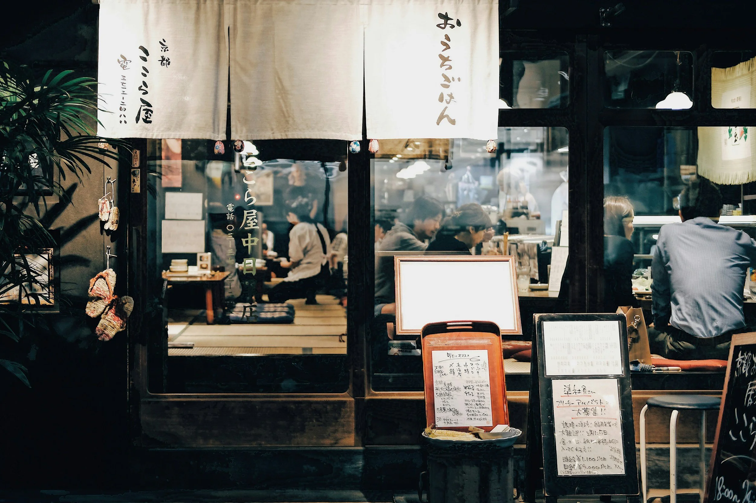 A Japanese restaurant front with a window showing customers dining inside. There are handwritten menu signs and decorative elements outside, including hanging cloth banners and potted plants.