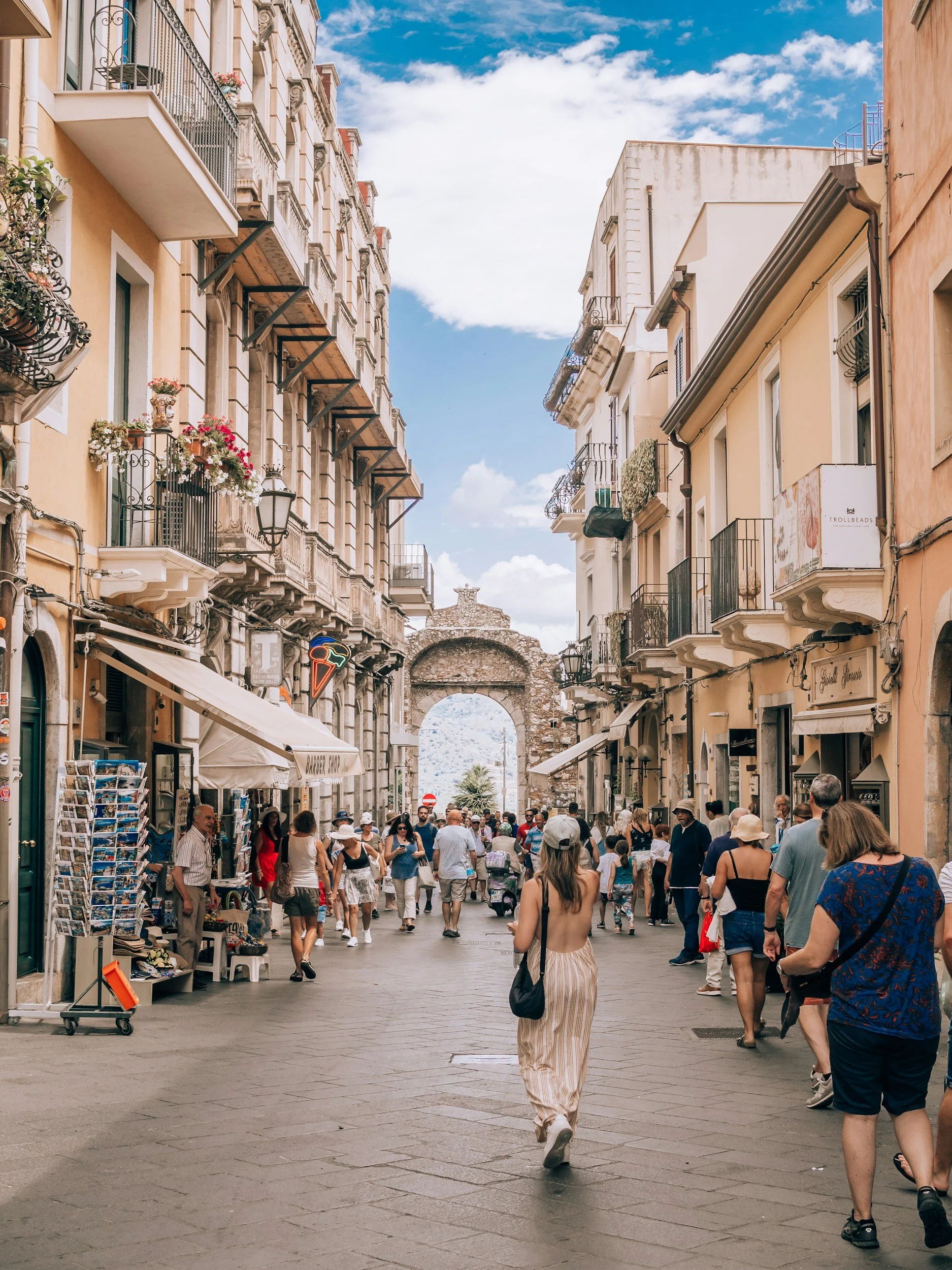 A busy street scene in a European city with pedestrians walking along the sidewalk. The street is lined with shops, cafes, and apartments with decorative balconies. An ancient stone archway is visible at the end of the street, and the sky is partly cloudy with blue patches.