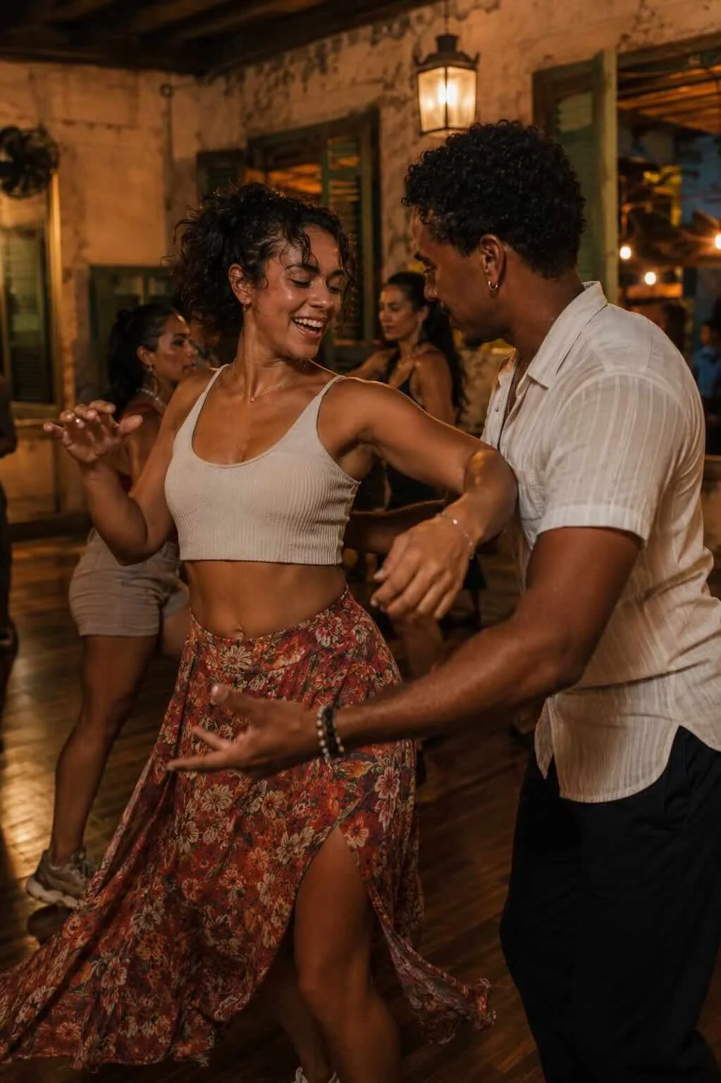 Couple dancing joyfully in a warmly lit indoor setting with other dancers in the background. The woman is wearing a beige crop top and a floral skirt, while the man is dressed in a light striped shirt and dark pants.