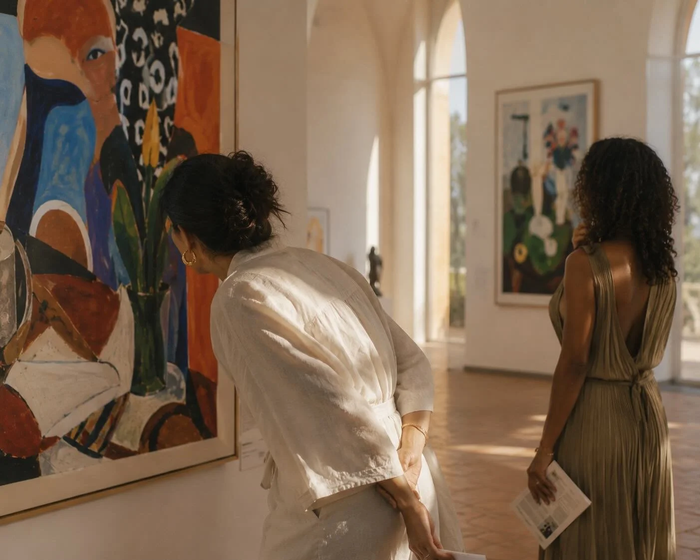 Two women in an art gallery observing paintings on the wall. One woman with curly dark hair and a gold dress is looking at a portrait, while the other woman with black hair tied back and in a white outfit is examining a colorful still life painting.