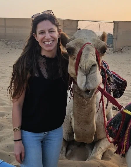 A woman smiling next to a camel with a red harness in a desert setting during sunset.
