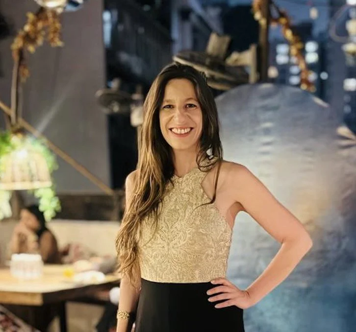 A woman in a sleeveless gold and black dress smiling with her hand on her hip, standing indoors with decorative lighting in the background.