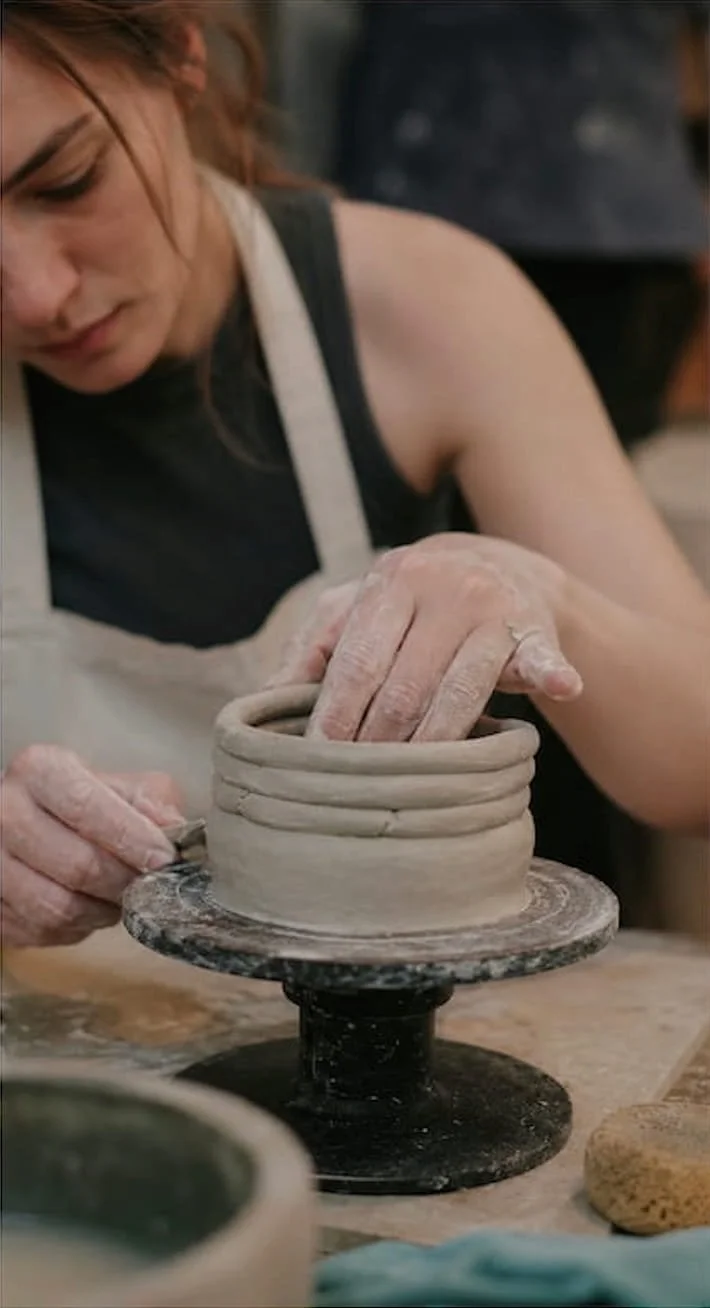 A woman shaping a clay pot on a rotating pottery wheel, wearing an apron, with her hands covered in clay.