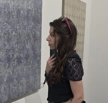 Side profile of a woman with long dark hair wearing sunglasses on her head, a black lace top, and holding a phone near her face, standing indoors against a textured wall.