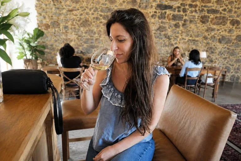 Woman with long dark hair sitting at a wooden table, holding a glass of white wine, in a cozy restaurant with an exposed stone wall, other patrons in the background.