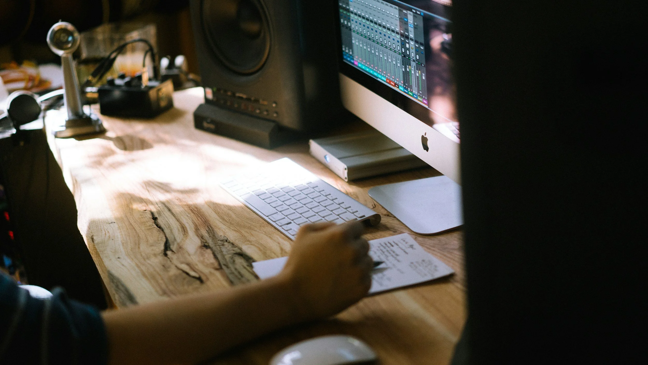 A computer workstation featuring an iMac with music production software on the screen, a wireless keyboard, and a person holding a piece of paper, set on a wooden desk with various electronic devices and black speakers in the background.