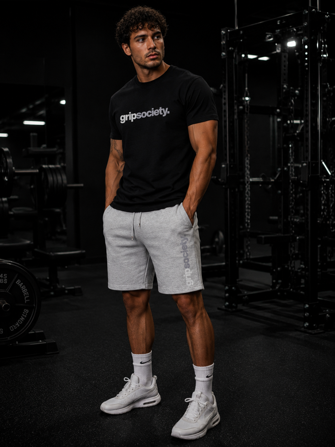 A young man standing in a gym, wearing a black t-shirt with 'gripsociety.' written on it, light gray shorts, white Nike sneakers, and white socks, with gym equipment in the background.