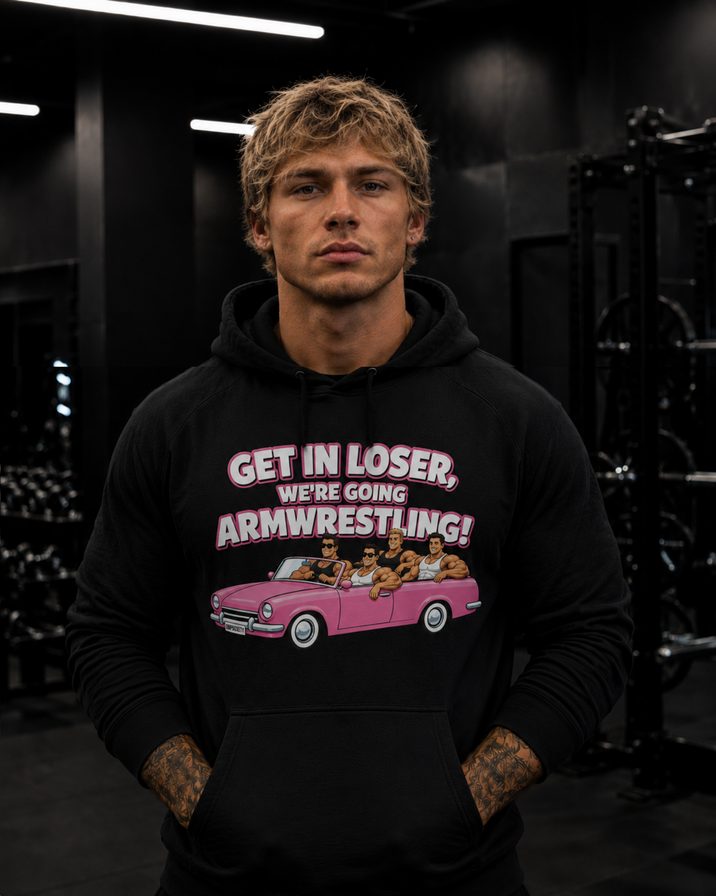 A young man with tattoos on his hands and blond curly hair, wearing a black hoodie with a graphic that says 'Get in loser, we're going armwrestling' and features animated characters in a pink car. He is standing in a gym with black walls and fitness equipment in the background.