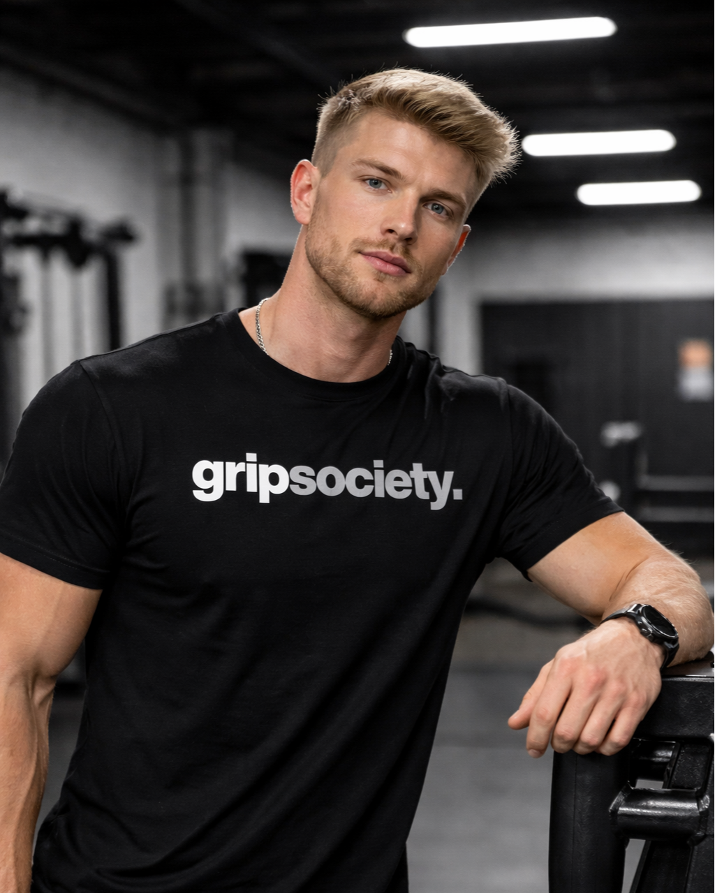 A young man with short blonde hair and a beard poses in a gym, wearing a black t-shirt with the words 'gripsociety.' and a black wristwatch, standing next to gym equipment with a confident expression.