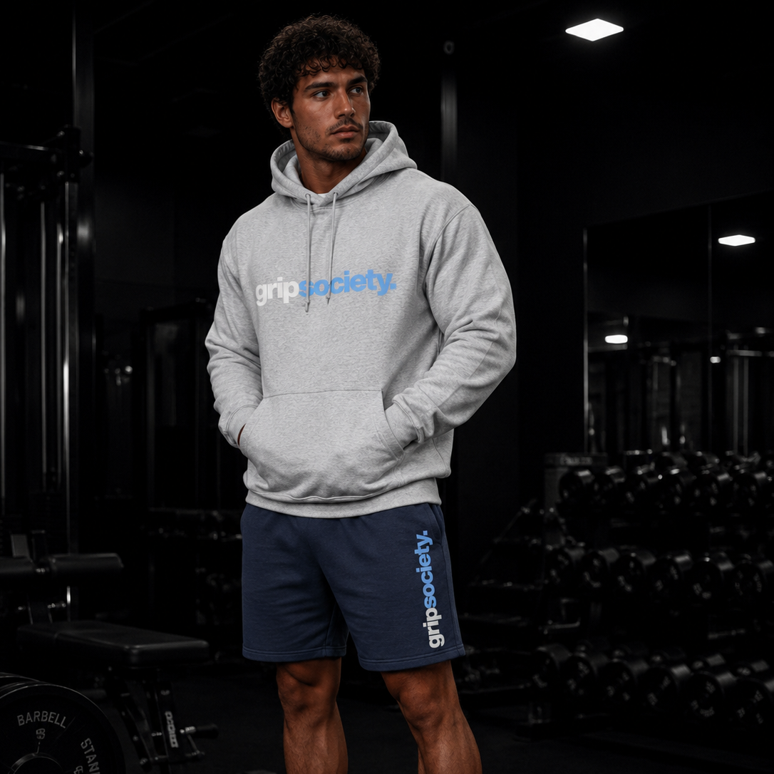 A young man in a gray hoodie and navy shorts with the words 'gripsociety' standing in a gym surrounded by black weights and equipment.