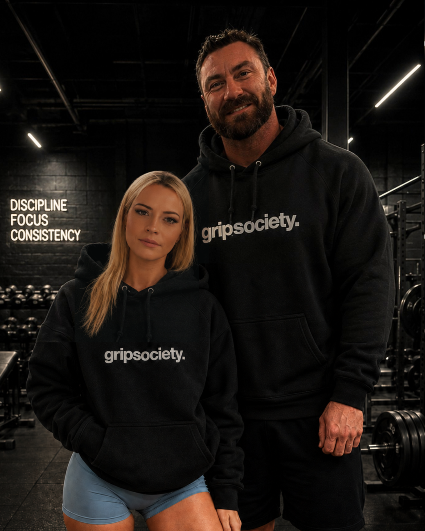 Two fitness enthusiasts in a gym wearing black hoodies with 'gripsocty.' written on them; a woman with blonde hair and a man with brown hair and a beard, both smiling. The background includes black brick walls, gym equipment, and motivational words 'Discipline Focus Consistency' on the wall.