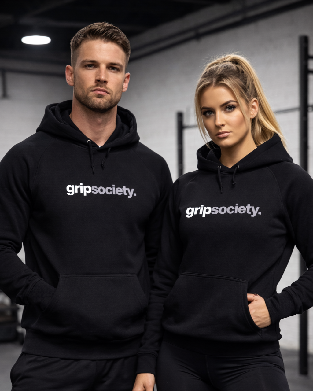 Two young adults, a man and a woman, standing in a gym wearing black hoodies with the logo 'gripsociety.'